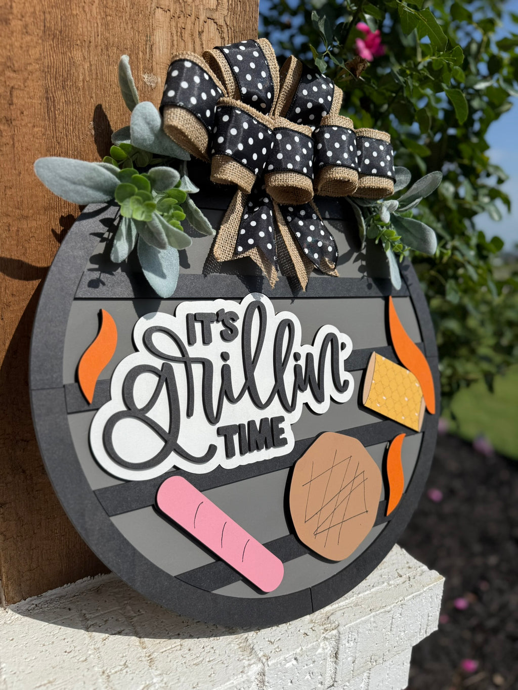 The Its Grillin Time Door Sign, Door Wreath hangs on a wooden post and features steak, hot dog, cheese, and flame accents plus a polka dot bow with greenery—making it the perfect decoration for your next BBQ.