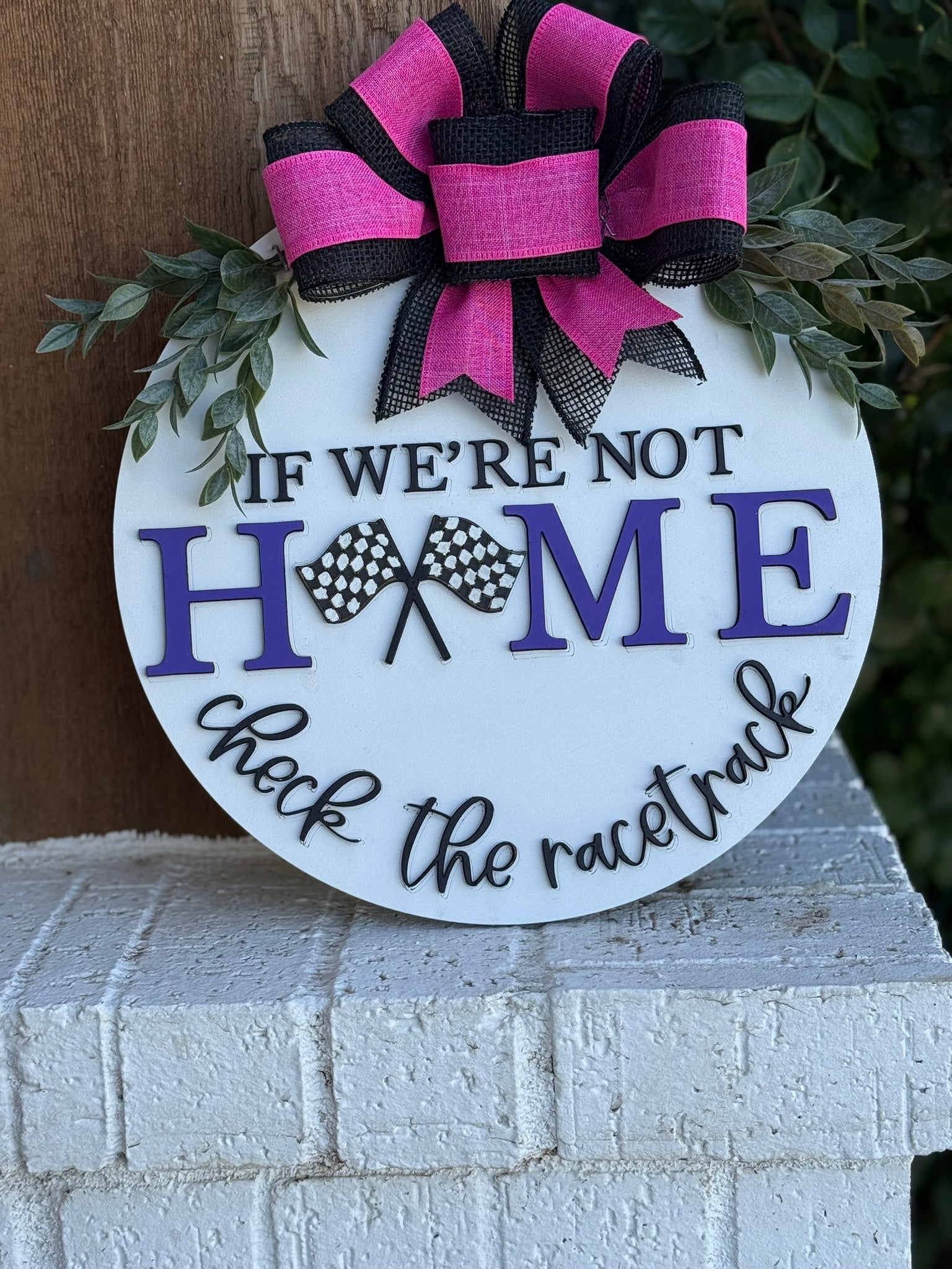 A round sign decorated with a pink and black bow and greenery reads, IF WERE NOT HOME check the racetrack, with HOME in purple letters and two checkered race flags in the center.