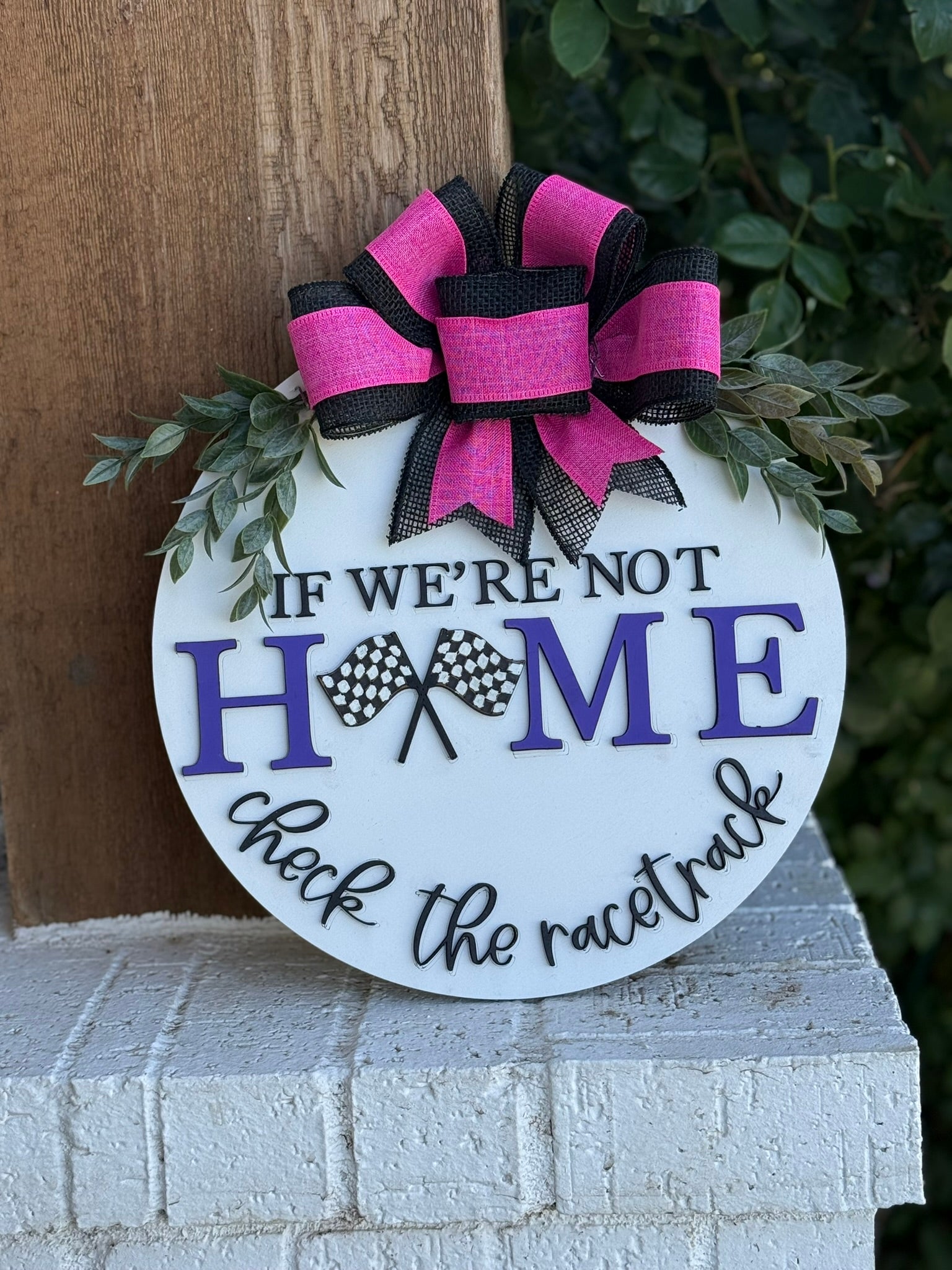 A round sign with a pink and black bow and greenery reads, If were not home, check the racetrack, with the O in home replaced by two crossed checkered flags. The sign rests against a brick ledge.