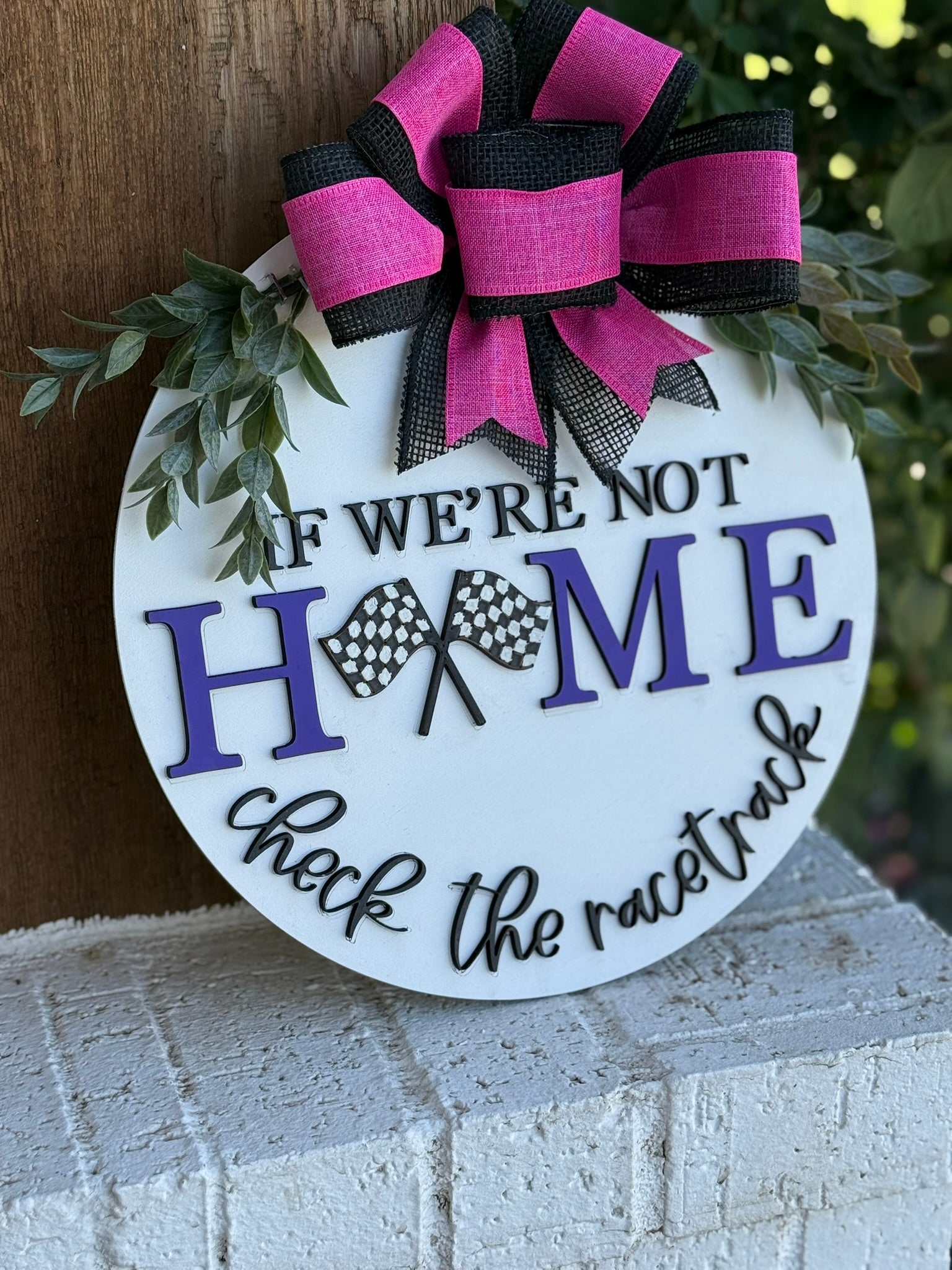 A round white sign with a pink and black bow and green leaves reads, If were not home, check the racetrack, with two checkered flags replacing the letter O in HOME. The sign is propped on a white brick surface.