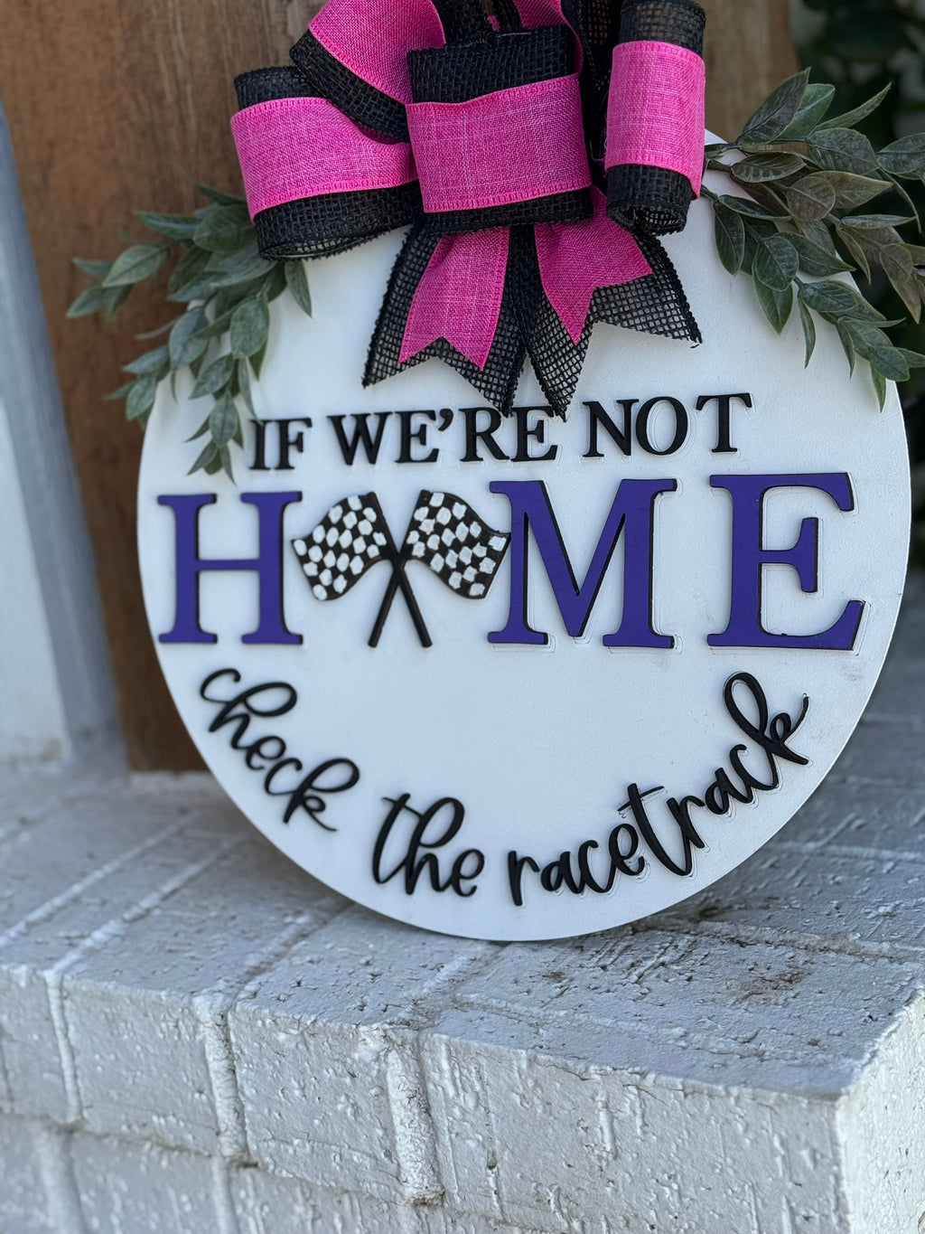 A round sign decorated with greenery and a pink-and-black bow reads, “If we’re not home, check the racetrack,” with the word “home” in purple letters and two checkered flags between the letters.