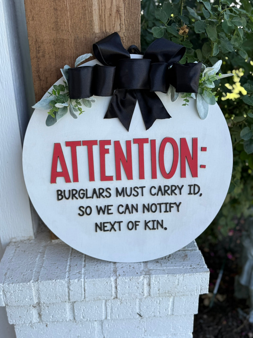 A round sign with “ATTENTION: BURGLARS MUST CARRY ID, SO WE CAN NOTIFY NEXT OF KIN” in bold letters, decorated with greenery and a large black ribbon, hanging on a porch column.