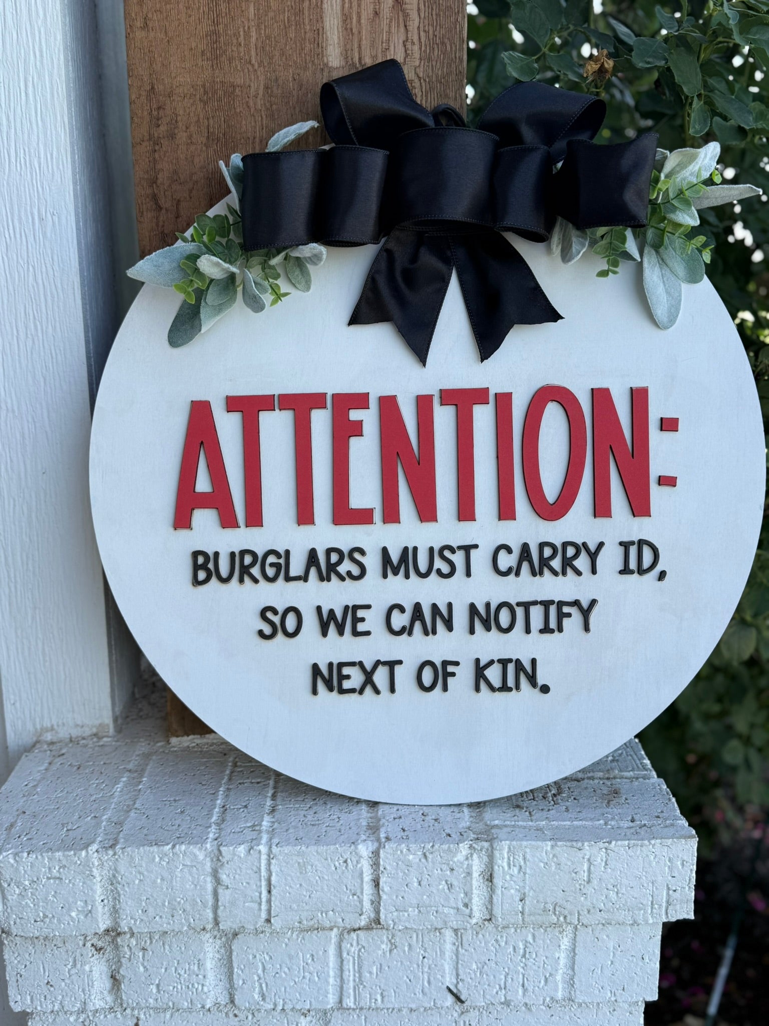 A round sign with a black bow and greenery reads: ATTENTION: BURGLARS MUST CARRY ID, SO WE CAN NOTIFY NEXT OF KIN. The sign is mounted on a white brick ledge near some greenery.