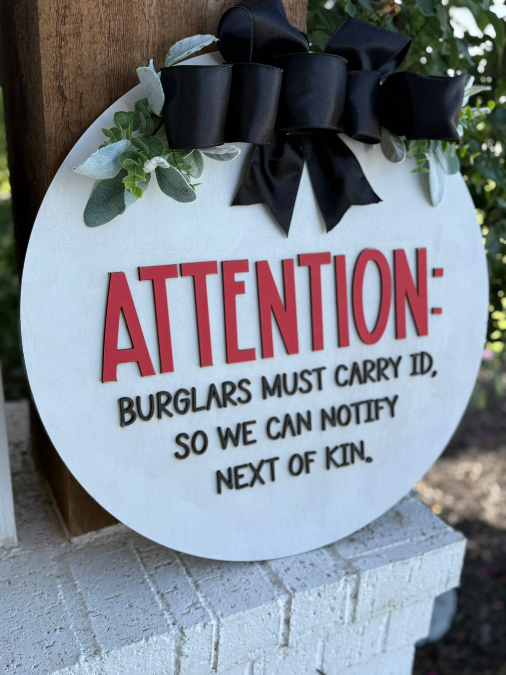 A round sign with a black bow reads: ATTENTION: BURGLARS MUST CARRY ID, SO WE CAN NOTIFY NEXT OF KIN. The sign is decorated with green leaves and hangs on a wooden post.