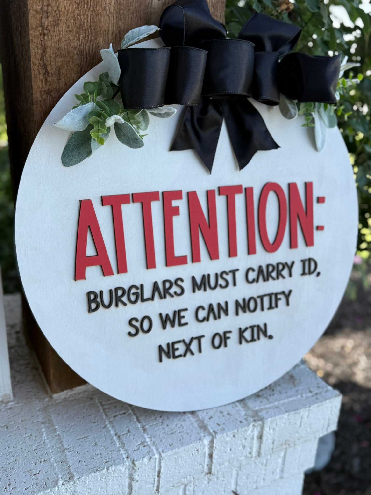 A round sign with a black bow reads: ATTENTION: BURGLARS MUST CARRY ID, SO WE CAN NOTIFY NEXT OF KIN. The sign is decorated with green leaves and hangs on a wooden post.