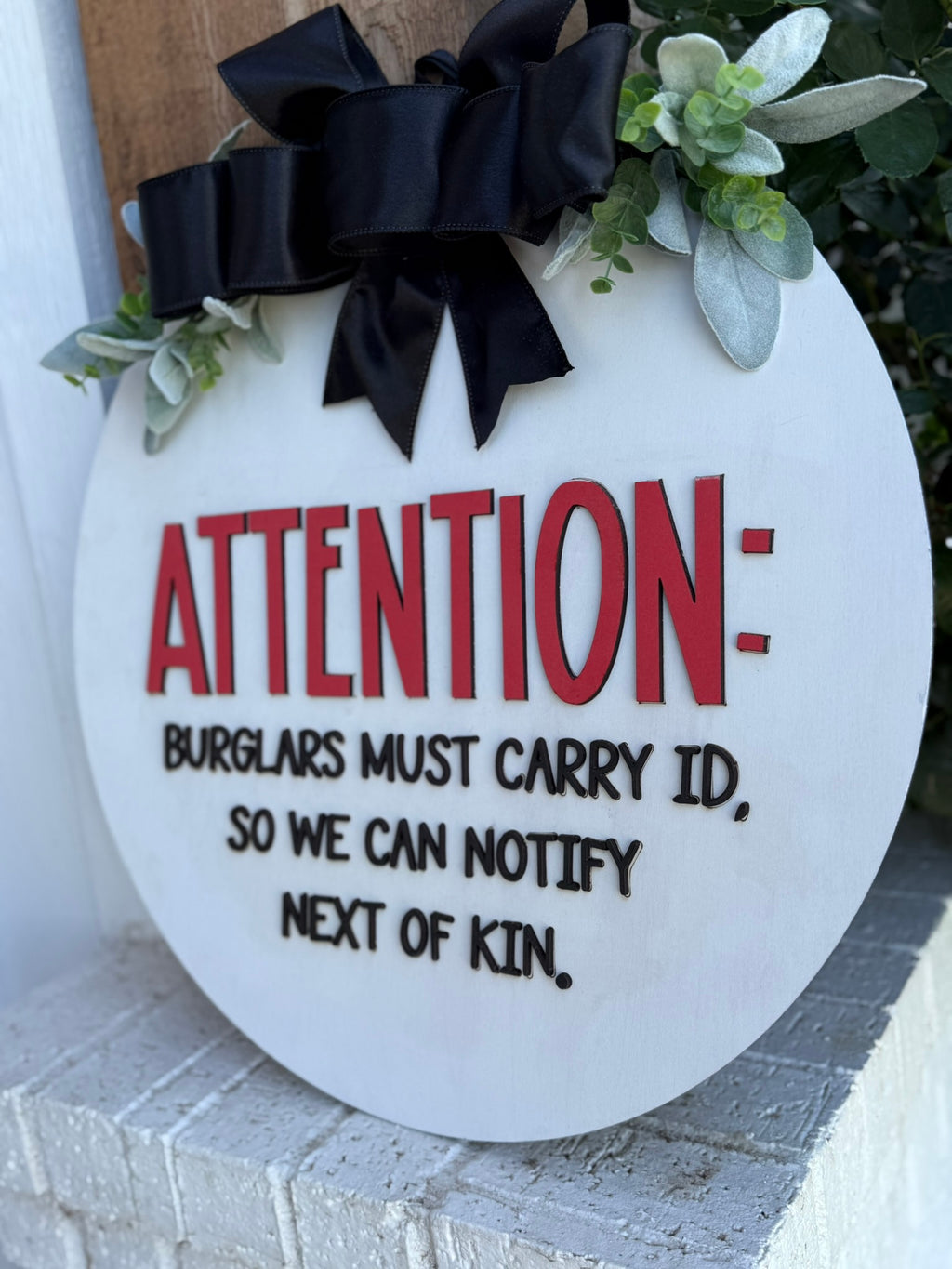 A round sign decorated with greenery and a black bow reads: ATTENTION: Burglars must carry ID, so we can notify next of kin. The text is in red and black on a white background, resting on a brick surface.
