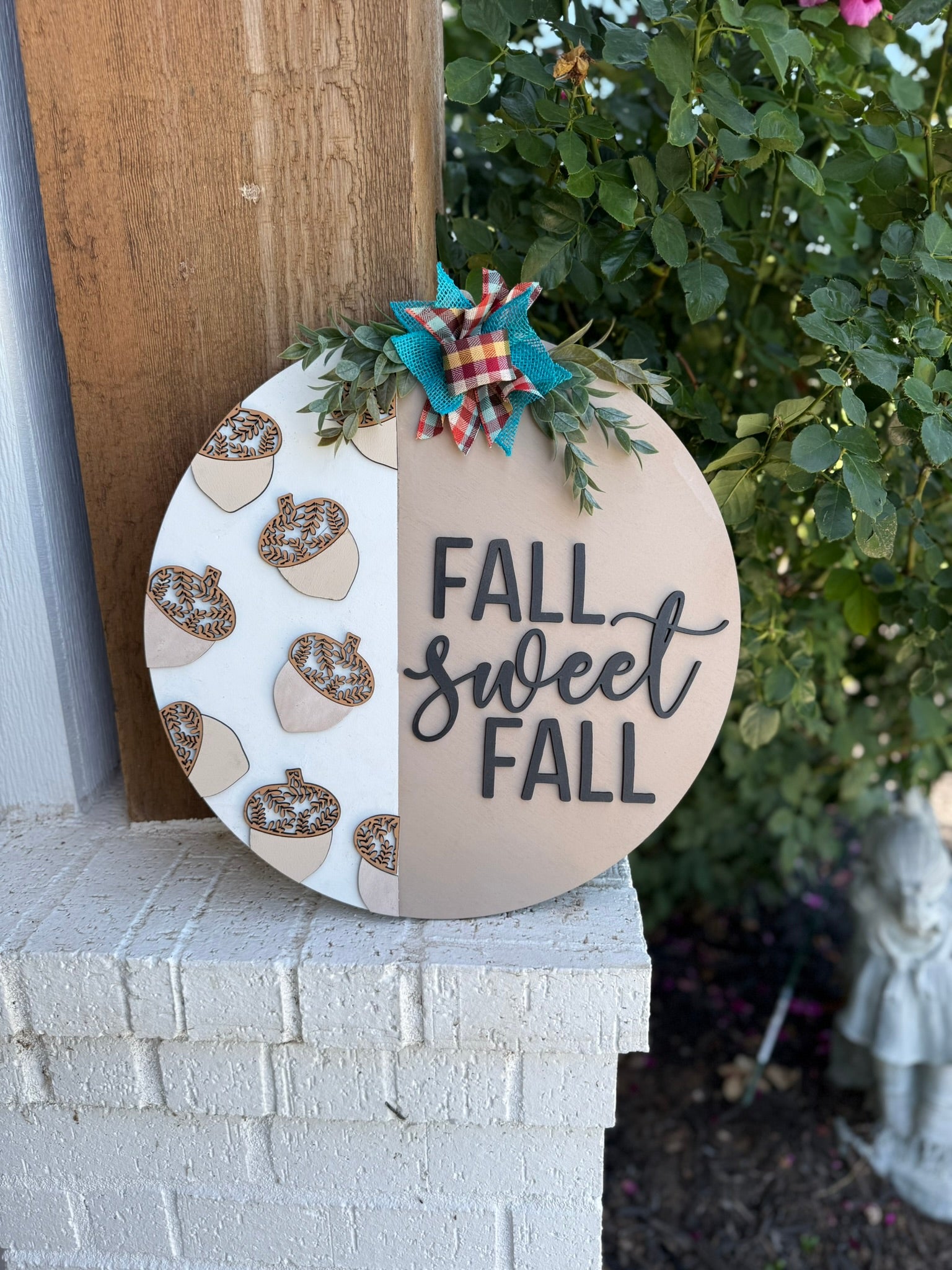 A round wooden sign on a brick ledge reads “FALL SWEET FALL.” It’s decorated with acorns on one side, a colorful plaid bow, greenery, and is placed next to a bush with greenery in the background.