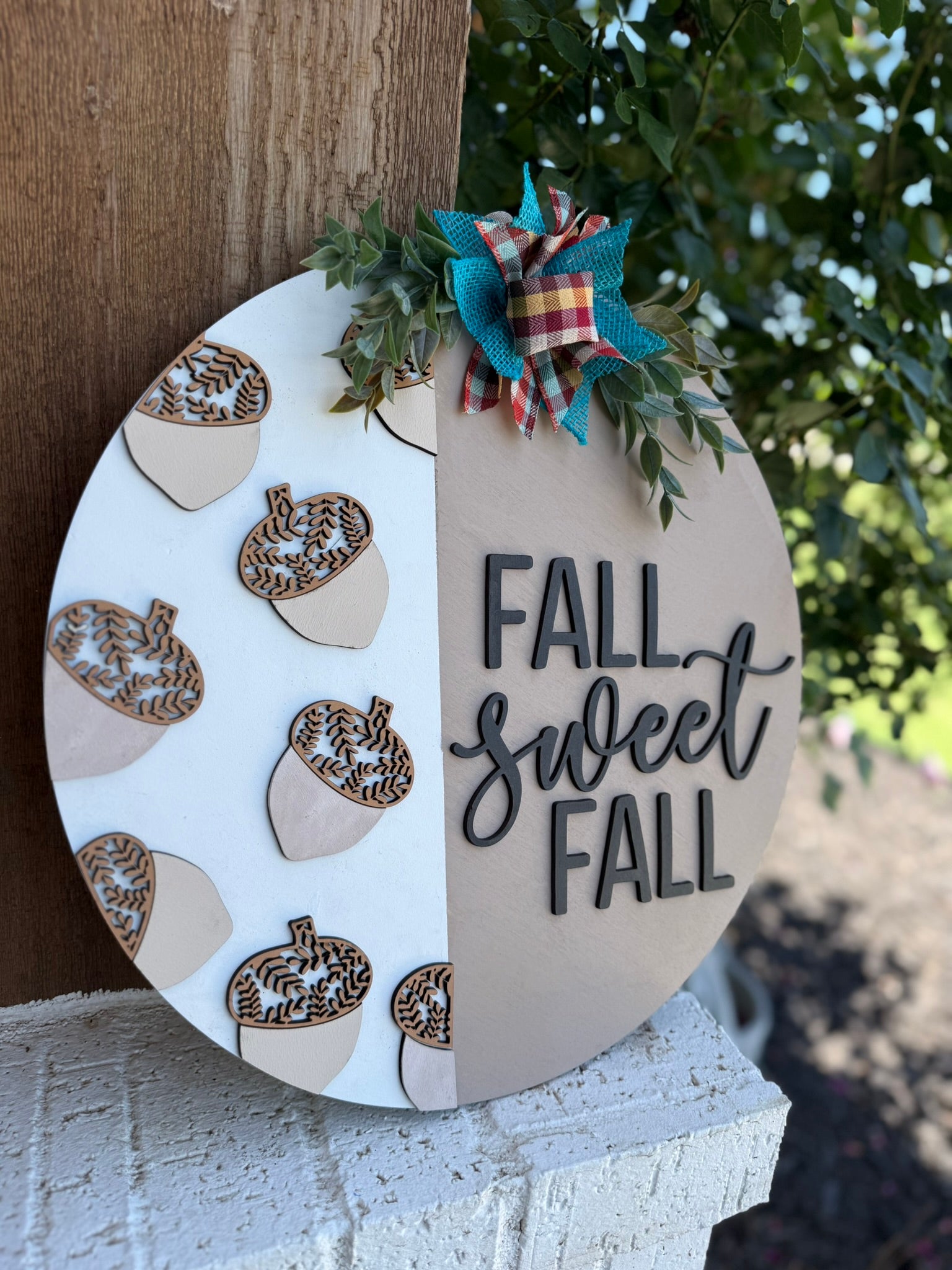 A round decorative sign with acorn accents, a plaid and teal bow, greenery, and the phrase “FALL SWEET FALL” displayed outdoors against a wooden wall and greenery.