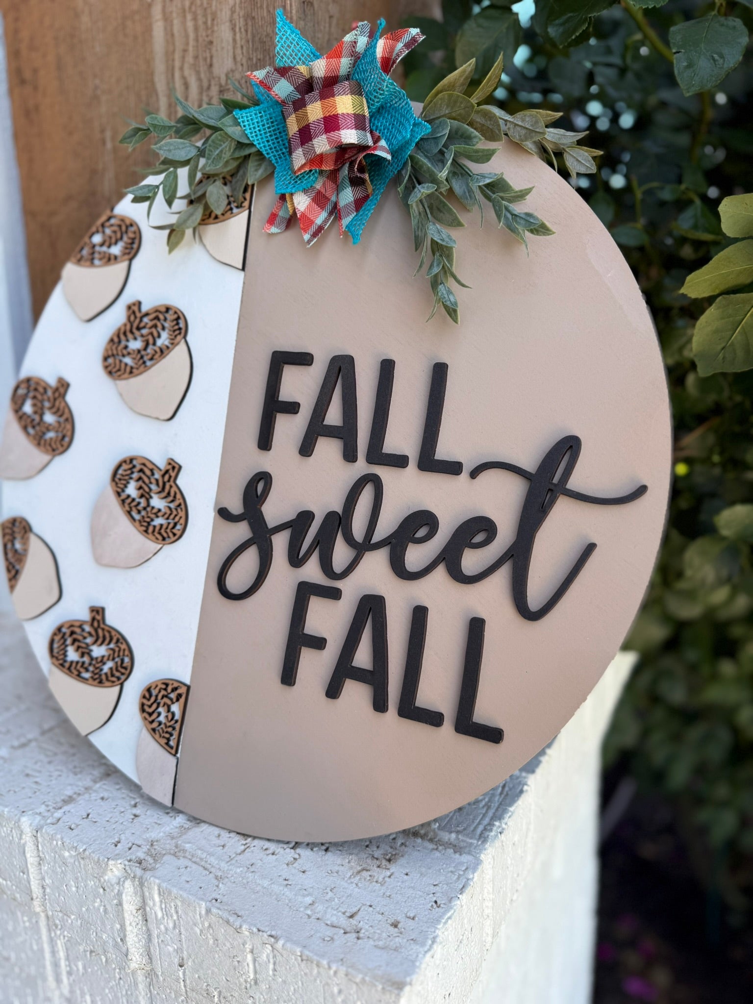 A round sign decorated with a bow and greenery reads FALL sweet FALL. One half has pumpkin illustrations, while the other half is plain with the text. The sign is displayed outdoors near plants.