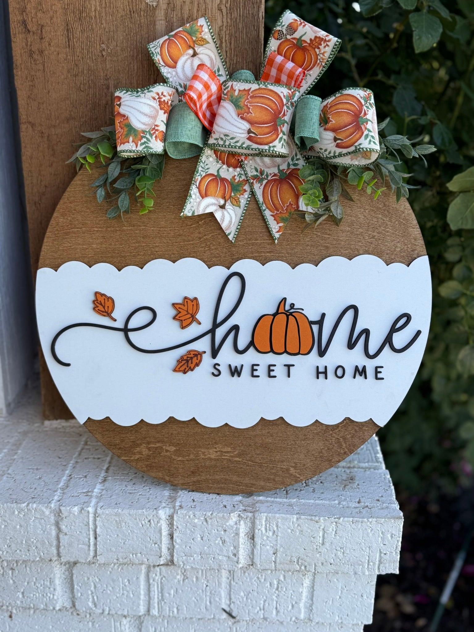 A round wooden sign with a white scalloped section reads Home Sweet Home, with a pumpkin replacing the o in home. It’s decorated with a fall-themed bow and greenery at the top.