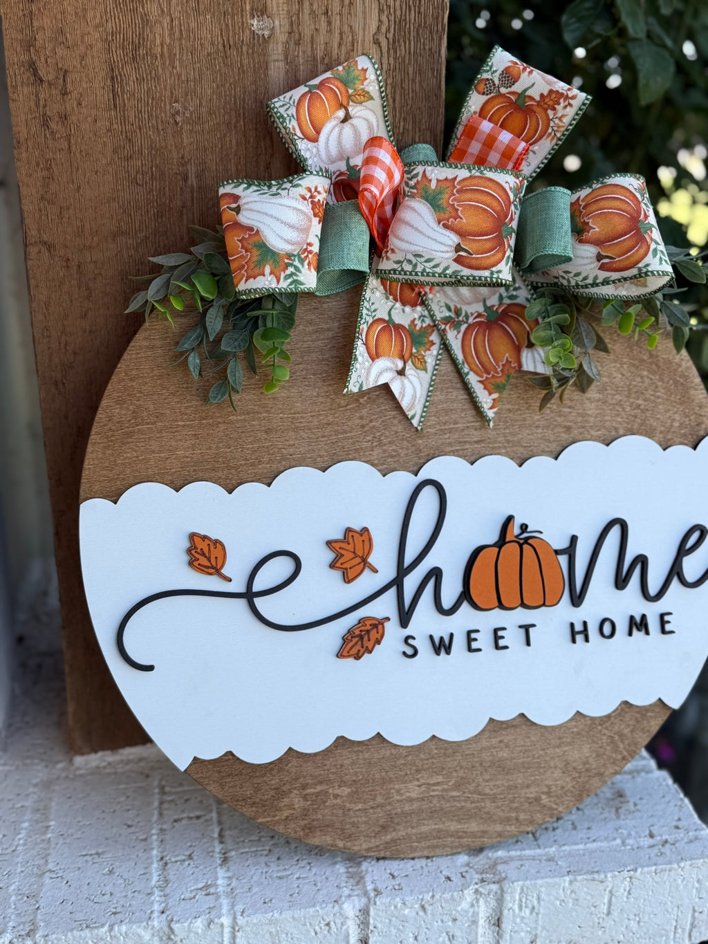 A round wooden sign decorated with fall leaves, a pumpkin, and a bow featuring pumpkins and green ribbon. The sign reads “Home Sweet Home,” with the “o” in “Home” replaced by a pumpkin.