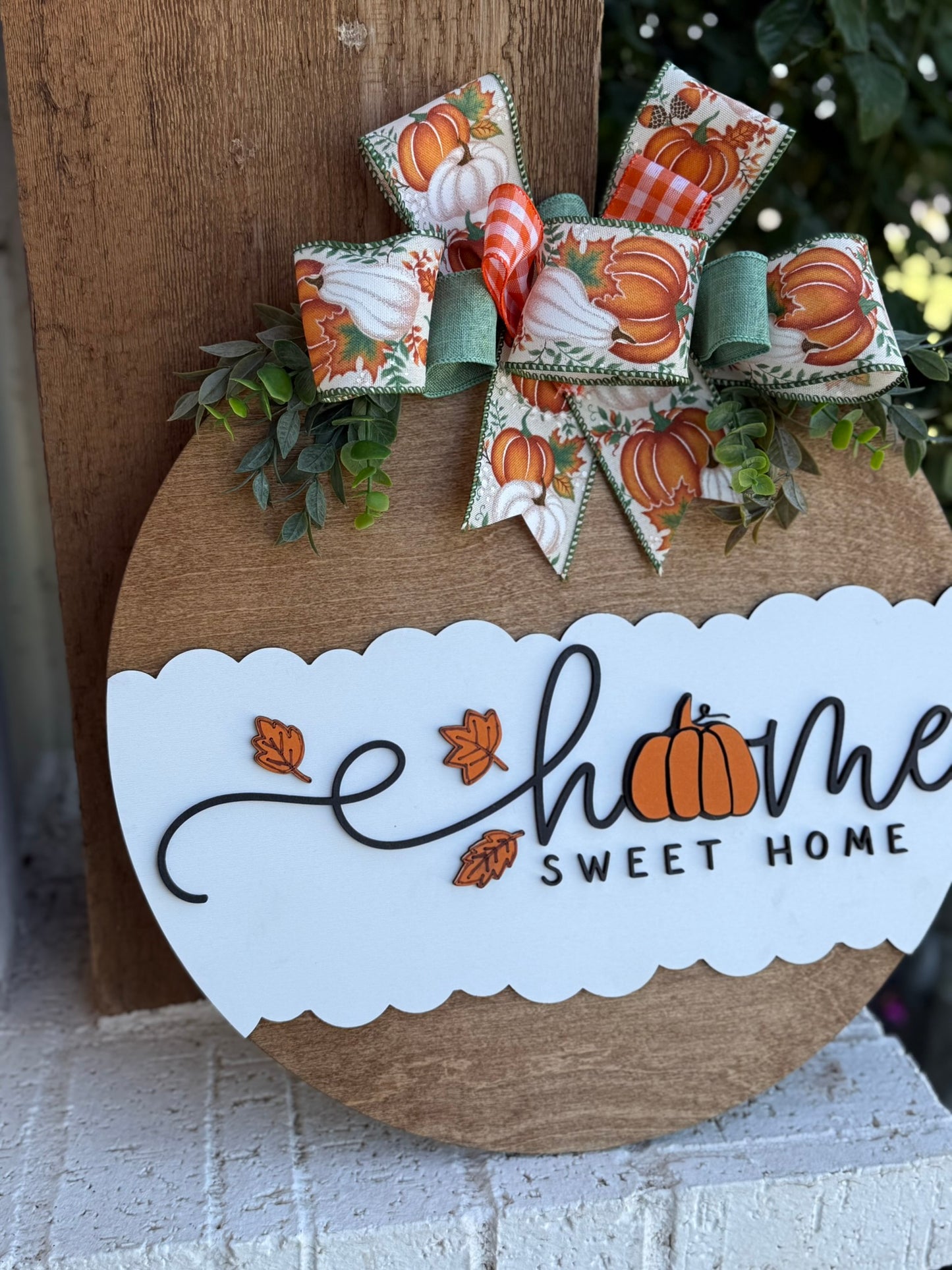A round wooden sign decorated with fall leaves, a pumpkin, and a bow featuring pumpkins and green ribbon. The sign reads “Home Sweet Home,” with the “o” in “Home” replaced by a pumpkin.