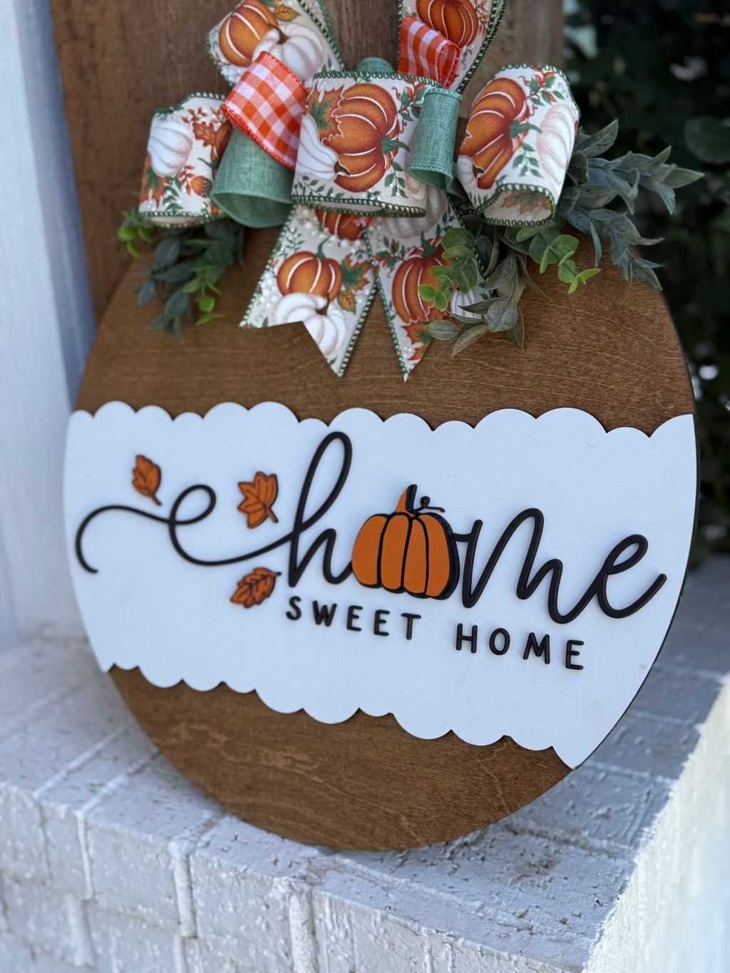 A round wooden sign with a white scalloped section reads Home Sweet Home, with a pumpkin replacing the o in home. It’s decorated with a fall-themed bow and greenery at the top.