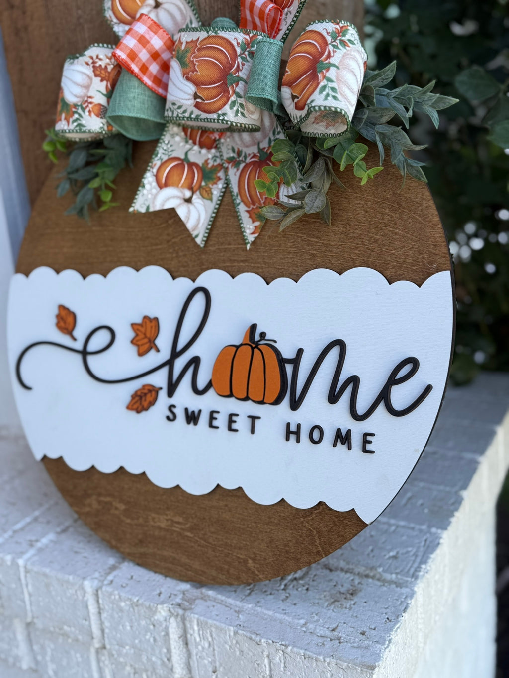 A round wooden sign with a white scalloped section reads Home Sweet Home, with a pumpkin replacing the o in home. It’s decorated with a fall-themed bow and greenery at the top.