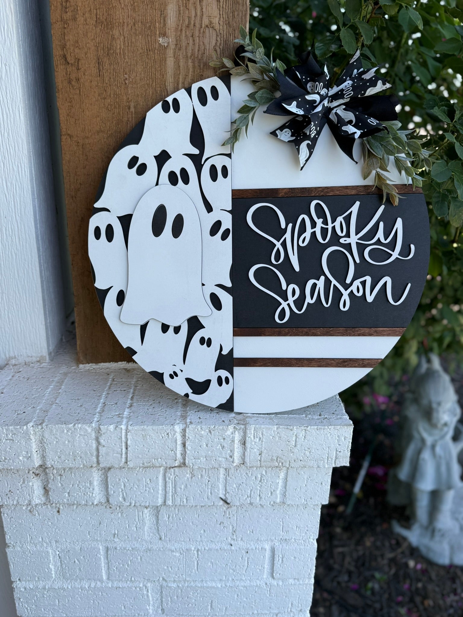 A round wooden sign with Spooky Season written on one half and a pattern of white cartoon ghosts on the other, decorated with a black and white ribbon and greenery, leaning on a white brick surface outdoors.