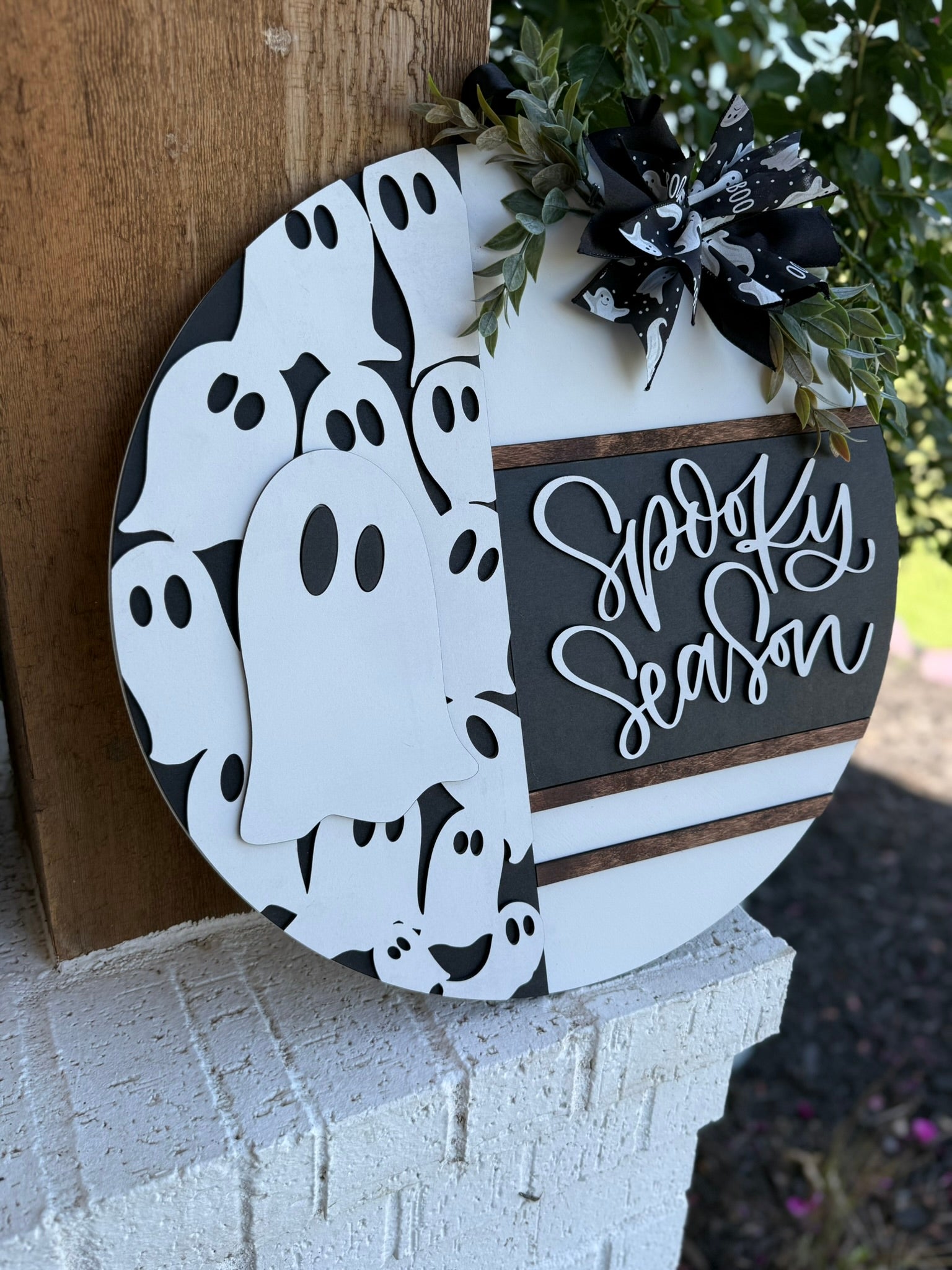 A round wooden sign with Spooky Season written on one half and a pattern of white cartoon ghosts on the other, decorated with a black and white ribbon and greenery, leaning on a white brick surface outdoors.