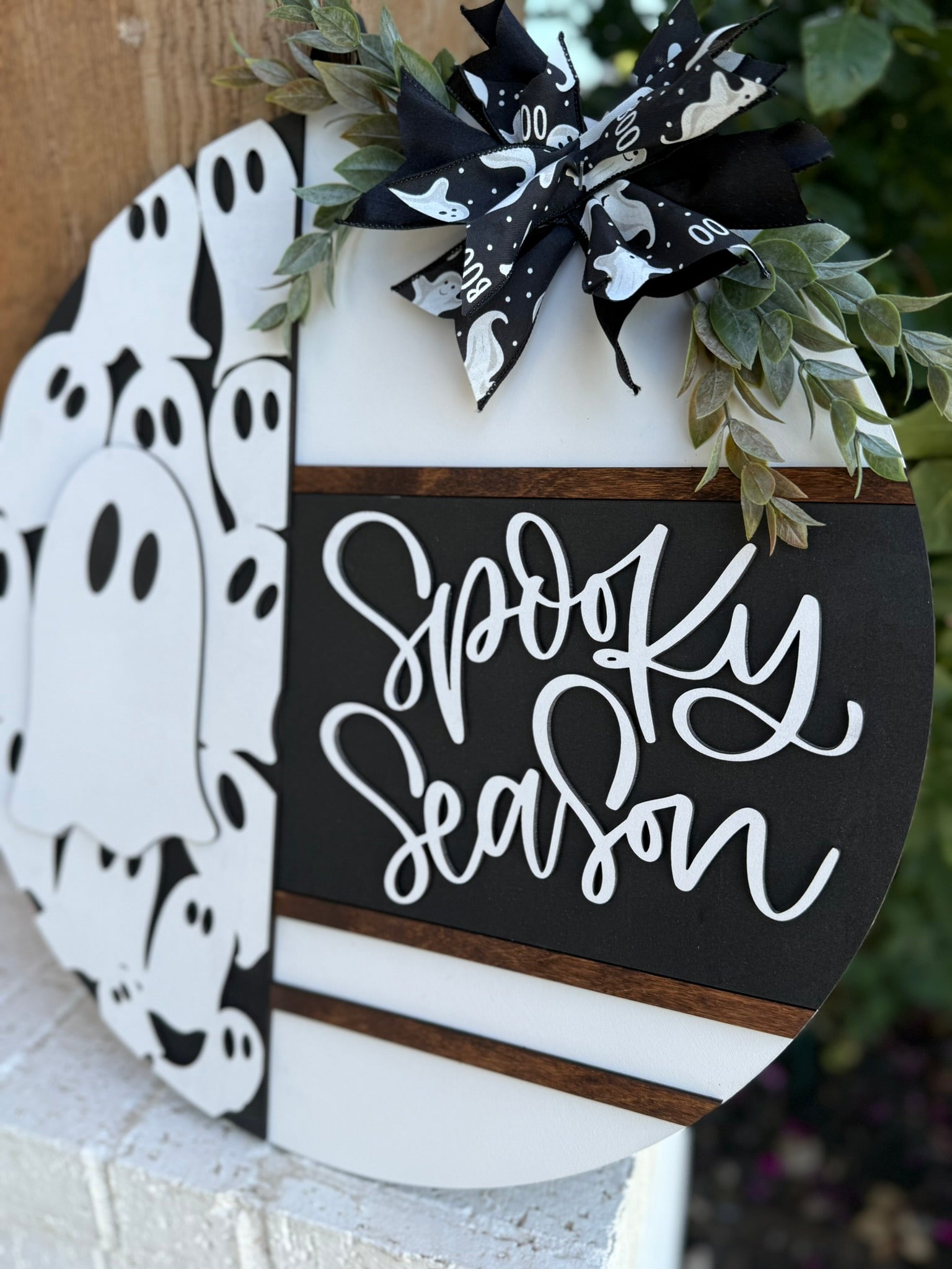 A round wooden sign with Spooky Season written on one half and a pattern of white cartoon ghosts on the other, decorated with a black and white ribbon and greenery, leaning on a white brick surface outdoors.