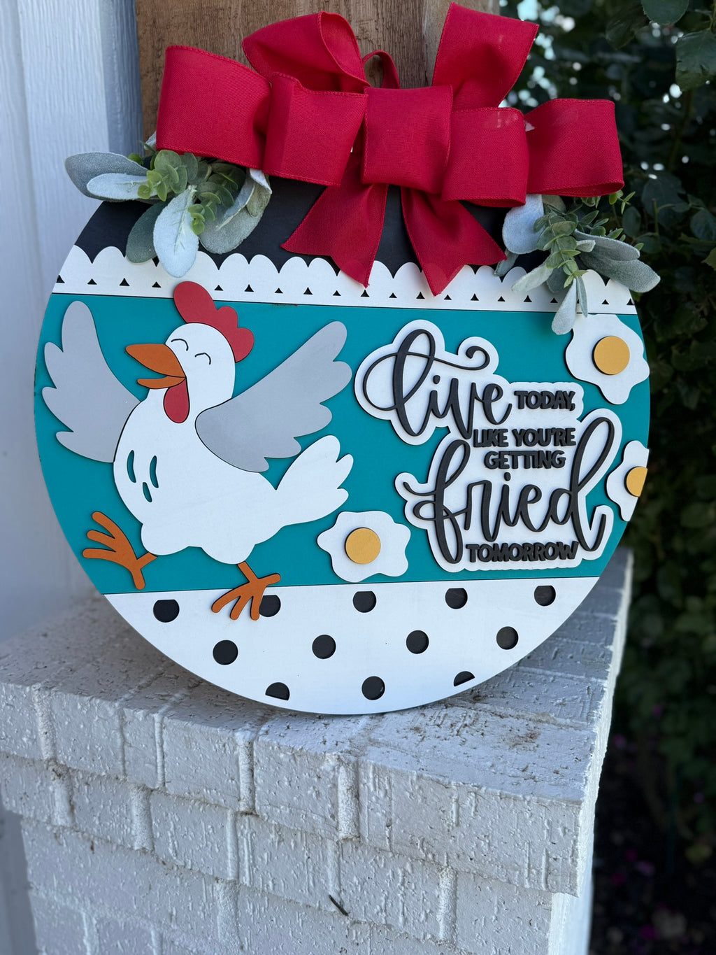 A round decorative sign with a cartoon chicken, eggs, and flowers, topped with red bows and greenery. The sign reads, Live today like youre getting fried tomorrow.