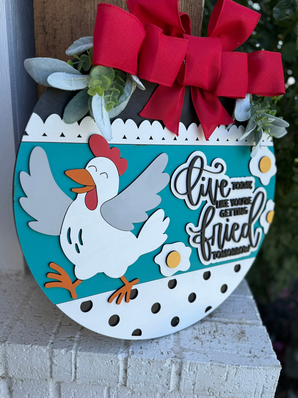 A round decorative sign with a cartoon chicken, fried eggs, a large red bow, and green leaves. Text reads, “Live today like you’re getting fried tomorrow.” The sign has polka dots and a teal background.