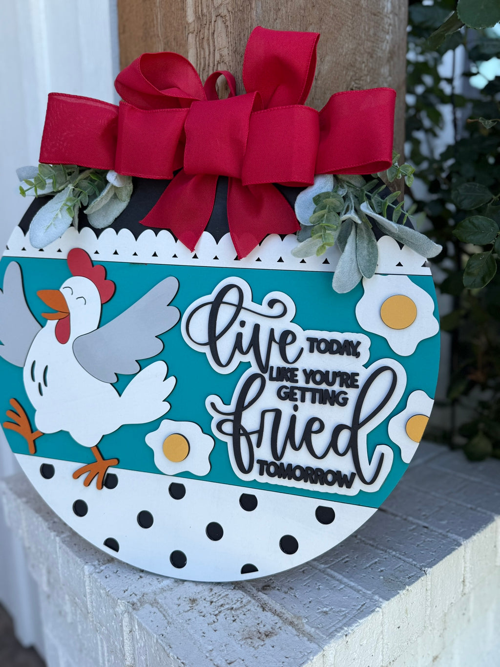A round decorative sign with a red bow, green leaves, a cartoon chicken, eggs, and black polka dots. The sign says, Live today like youre getting fried tomorrow.