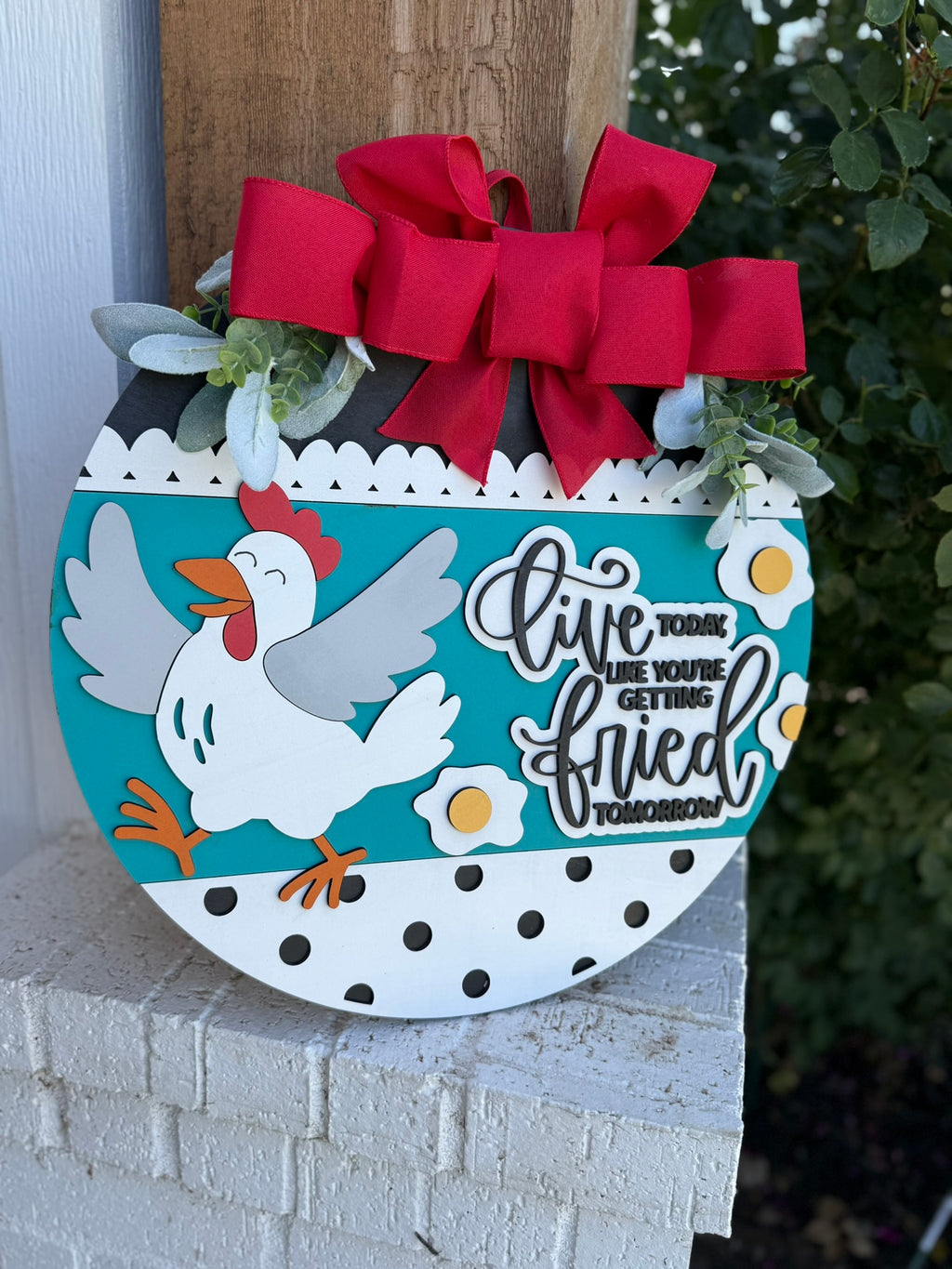 A round decorative sign with a cartoon chicken, a red bow, and greenery. The sign reads, Live today like youre getting fried tomorrow, with a polka dot border. The sign is displayed outdoors.
