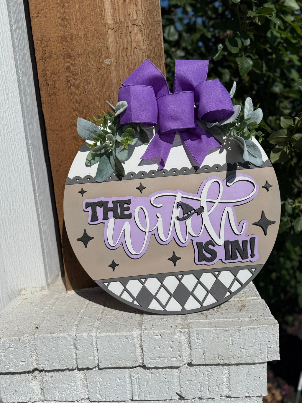 A round door sign decorated with a purple bow, greenery, and patterns reads The Witch is In! in bold and script letters, with a small witch silhouette flying on a broomstick.