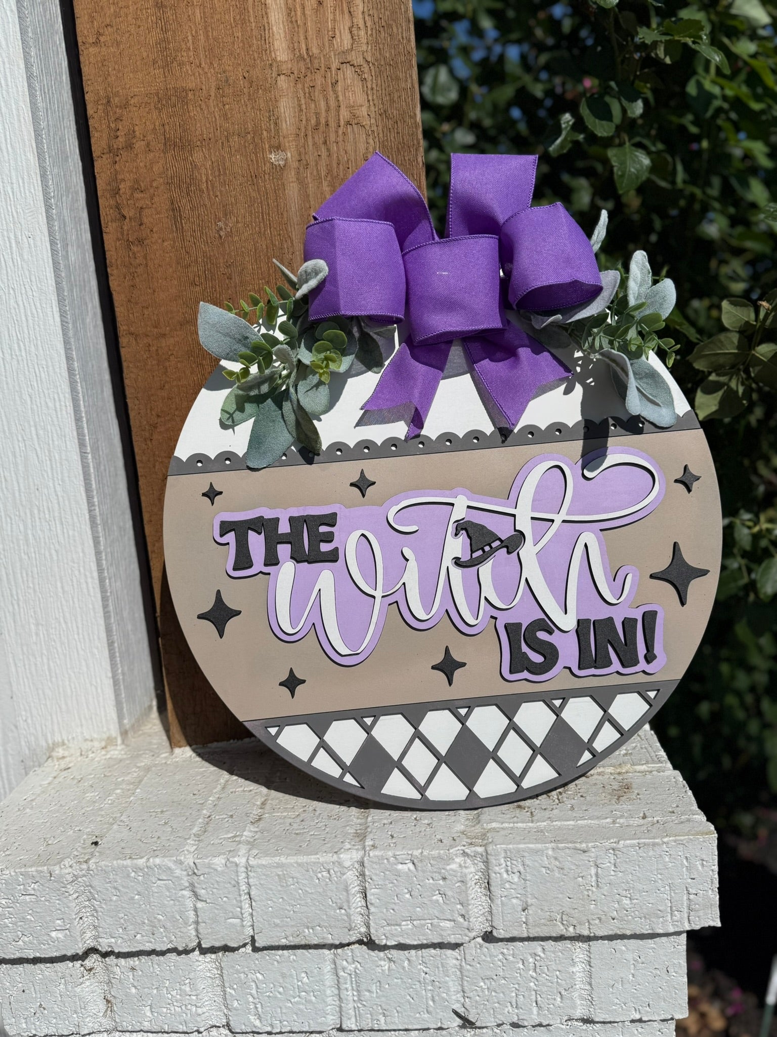 A round door sign decorated with a purple bow, greenery, and patterns reads The Witch is In! in bold and script letters, with a small witch silhouette flying on a broomstick.