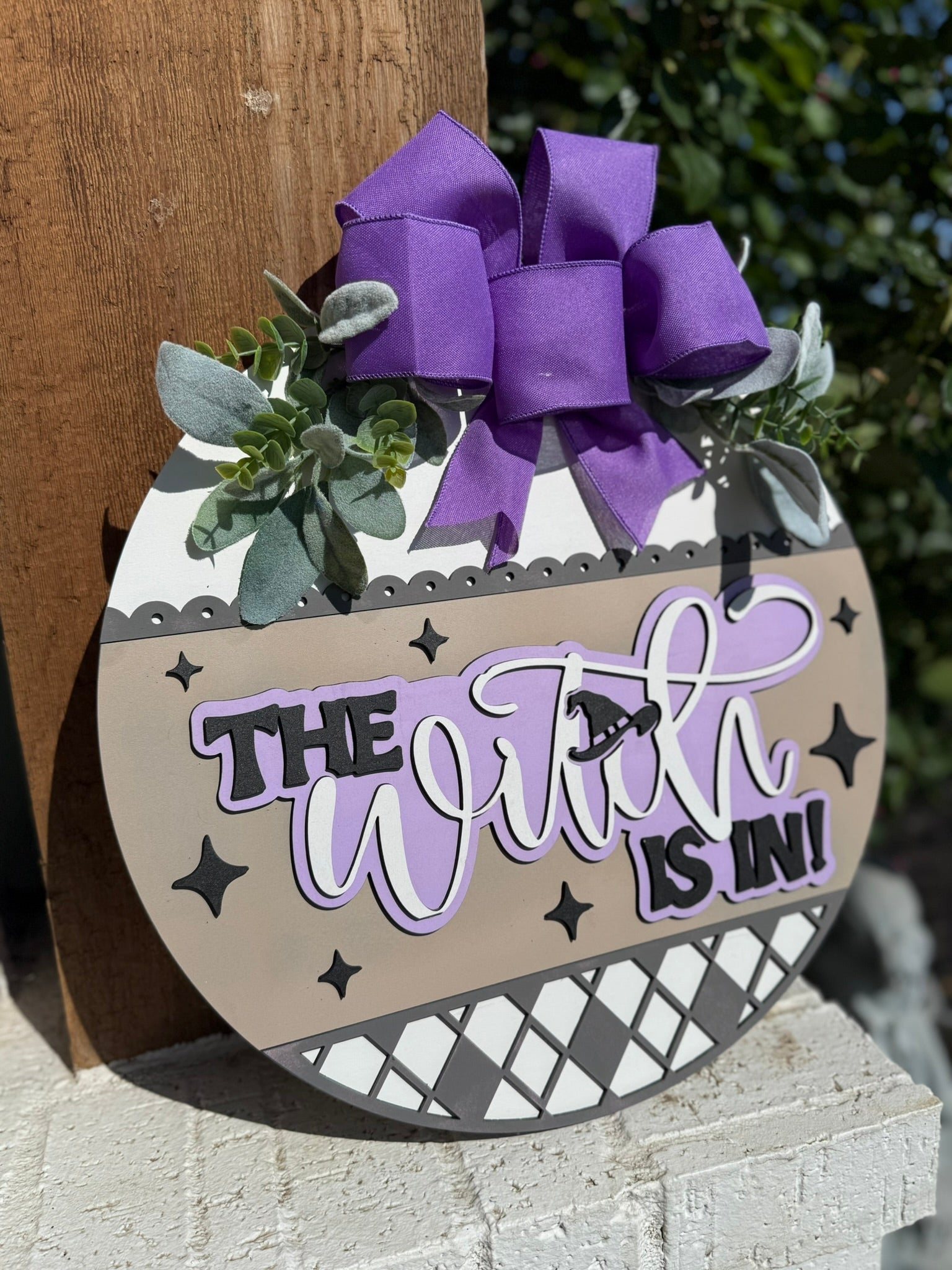 A round door sign decorated with greenery, a large purple bow, and the text The Witch Is In! in bold and script fonts, with a witch hat image and star accents. The sign has neutral tones and diamond patterns.