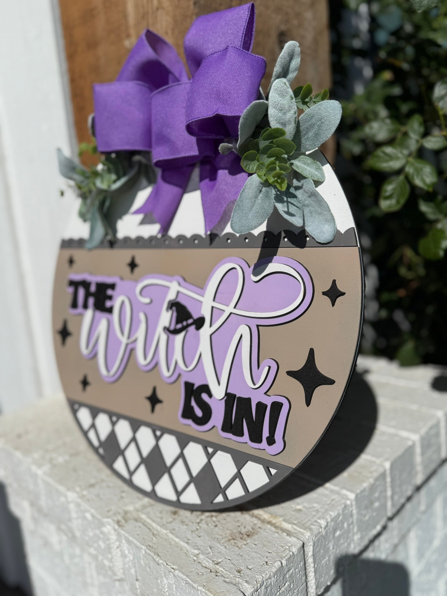 A round sign decorated with greenery, a large purple bow, and the phrase The Witch Is In! in bold, playful lettering, is displayed outdoors on a white brick surface.