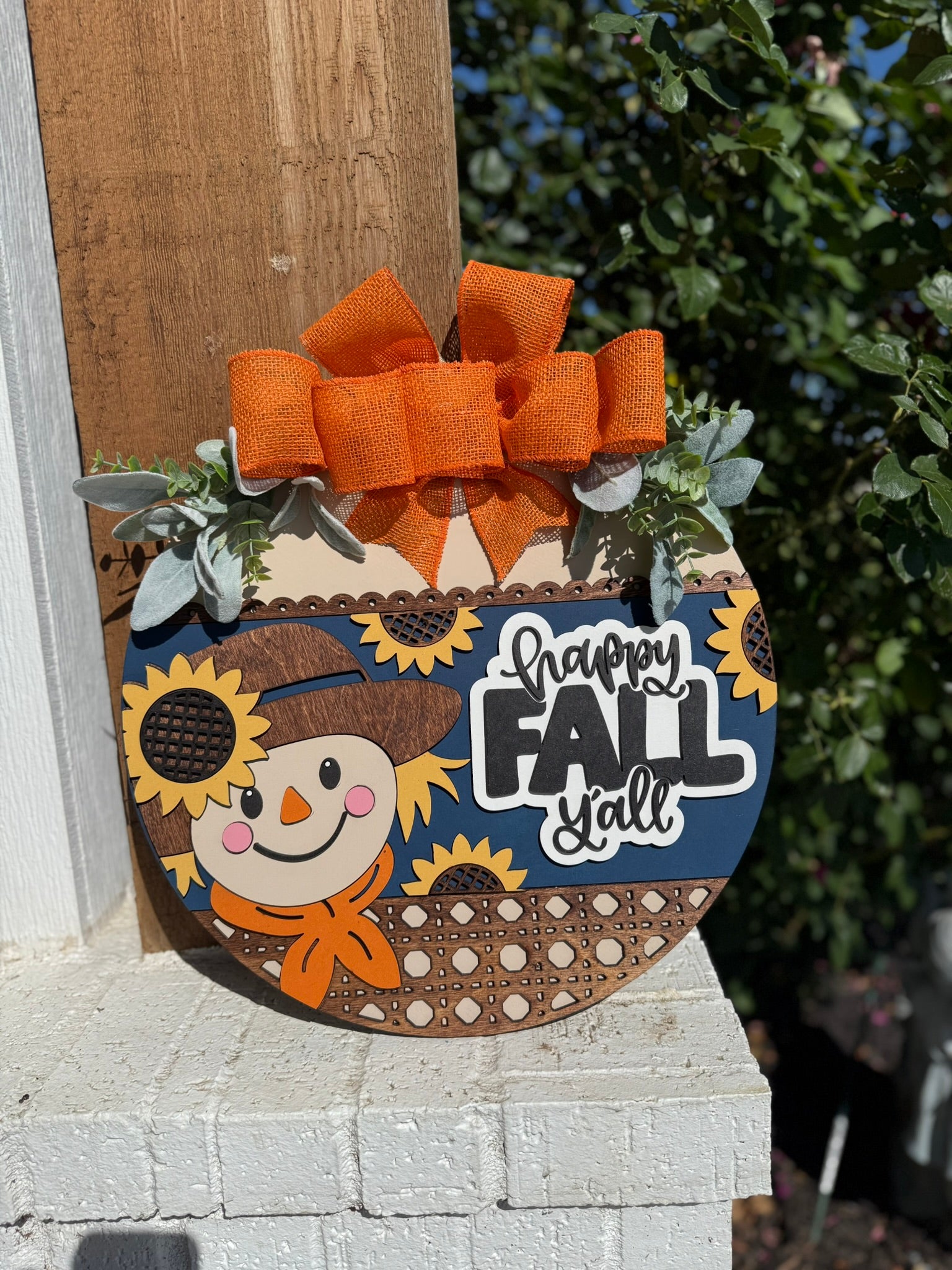A round wooden sign decorated with a smiling scarecrow, sunflowers, green leaves, and a large orange bow reads Happy Fall Yall. The sign is displayed outdoors on a white brick surface.