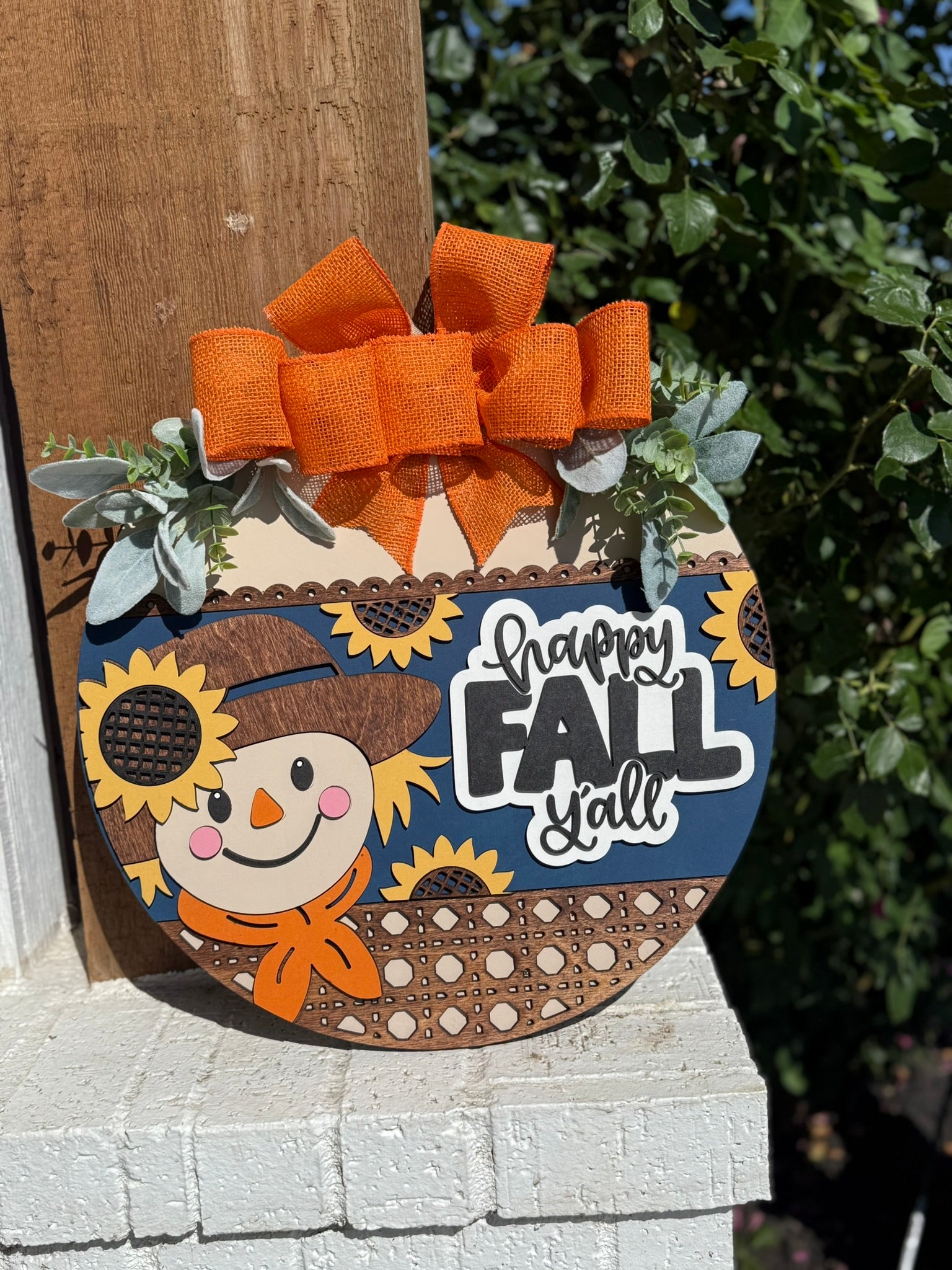 A round wooden sign with a scarecrow, sunflowers, and an orange bow reads “Happy Fall Y’all.” It is decorated with faux greenery and hangs on a post with greenery in the background.