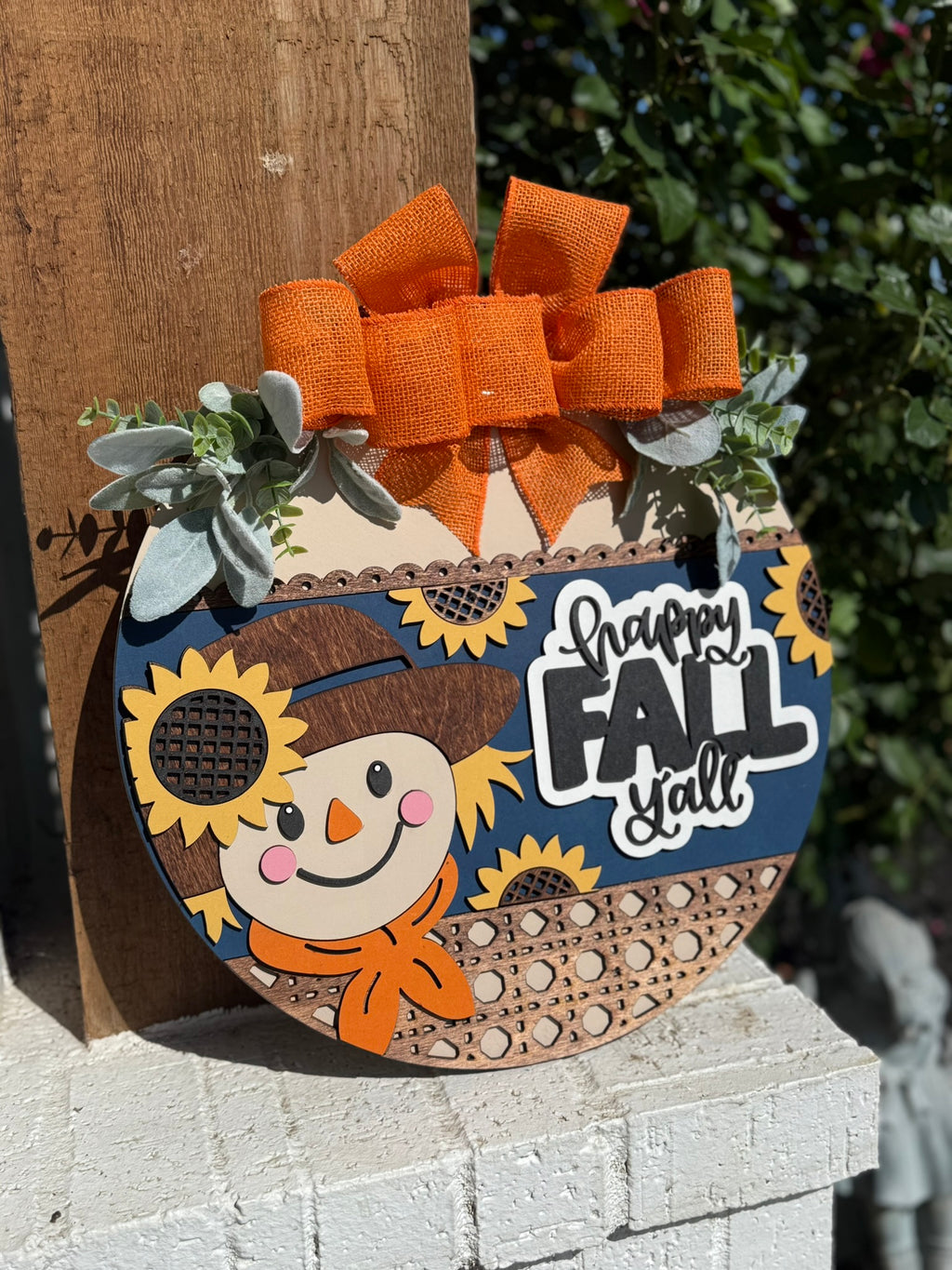 A decorative wooden sign shaped like a pumpkin features a smiling scarecrow, sunflowers, a large orange bow, greenery accents, and the words Happy Fall Yall in bold letters. The sign is set against an outdoor background.