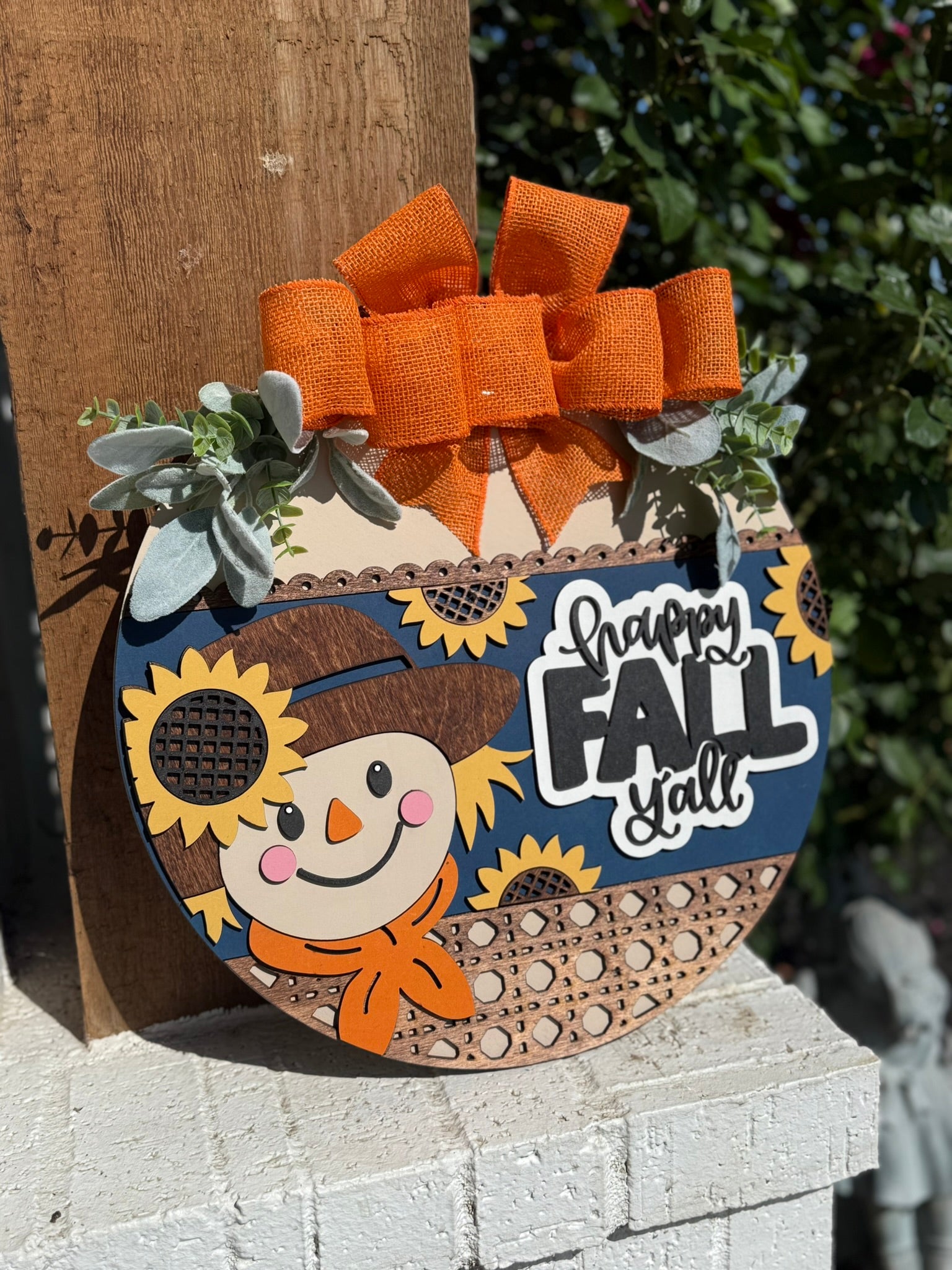 A decorative wooden sign shaped like a pumpkin features a smiling scarecrow, sunflowers, a large orange bow, greenery accents, and the words Happy Fall Yall in bold letters. The sign is set against an outdoor background.