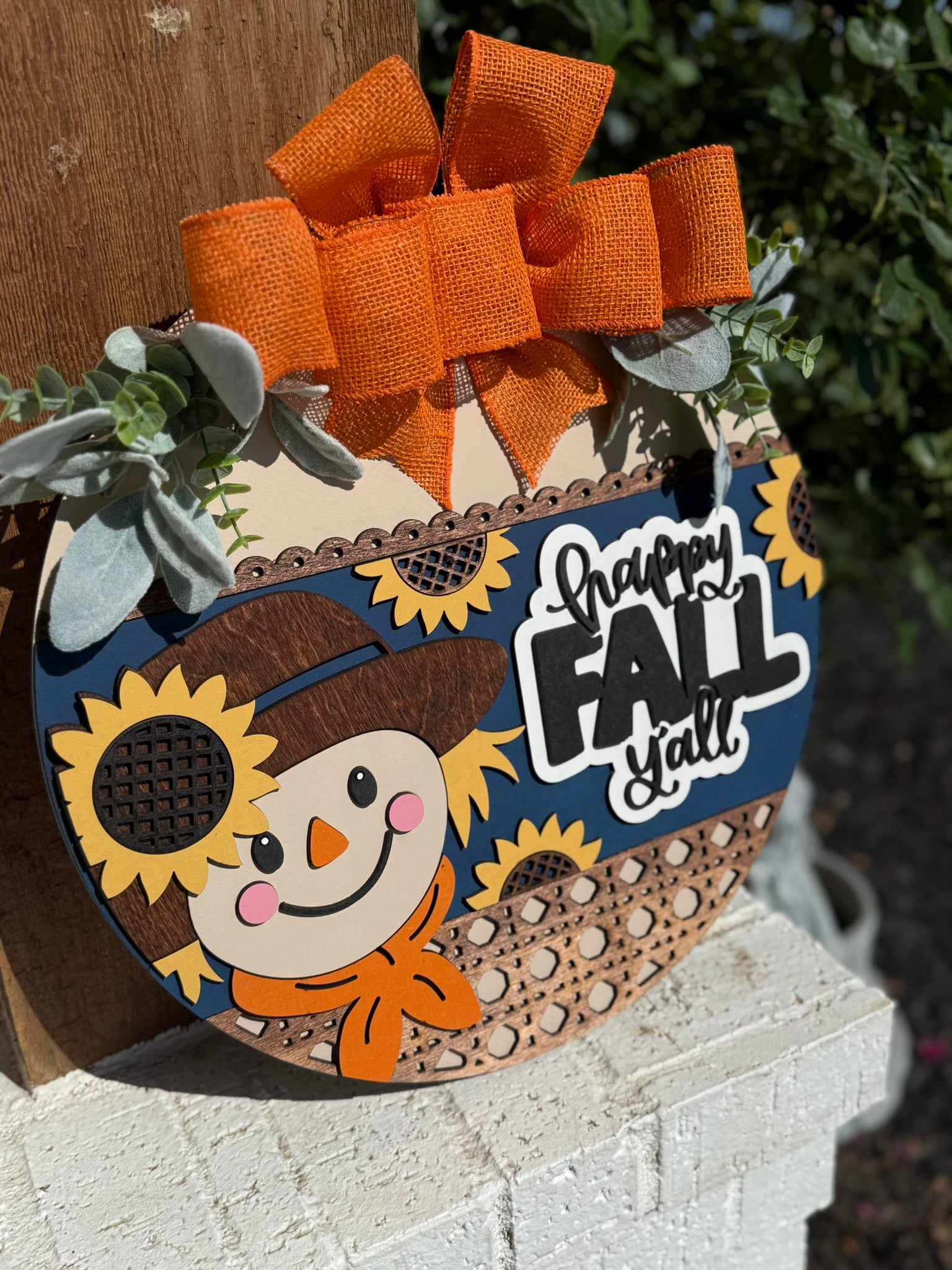 A round wooden sign decorated with a smiling scarecrow, sunflowers, greenery, and an orange bow. The sign reads Happy Fall Yall and is displayed on a porch railing.