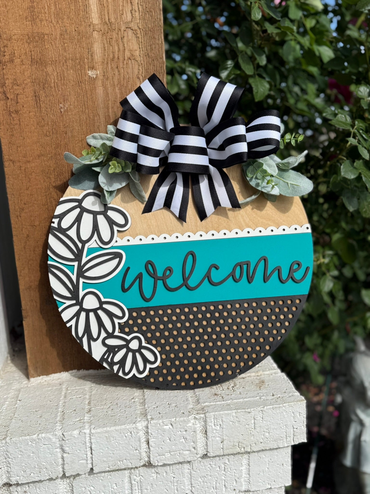 A round wooden welcome sign decorated with black and white flowers, polka dots, a teal strip with welcome written in cursive, leafy accents, and a large black-and-white striped bow at the top.