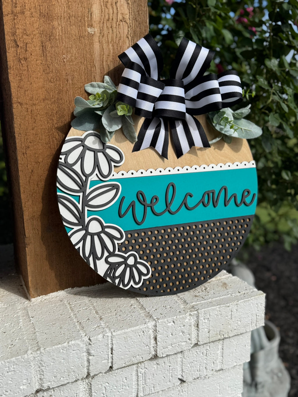 A round welcome sign with a striped black-and-white bow, artificial greenery, floral designs, and a teal stripe with the word “welcome,” hanging on a wooden post above a white brick surface.