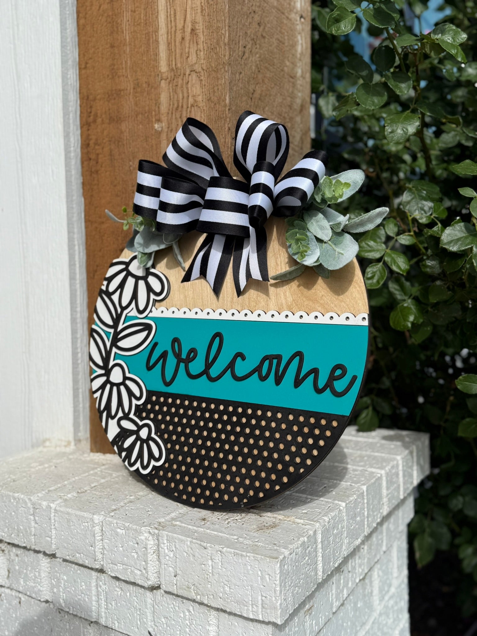 A decorative wooden sign with a black and white bow, faux greenery, and a painted design that includes the word welcome in teal, sits on a white brick ledge next to a wooden post and green bushes.