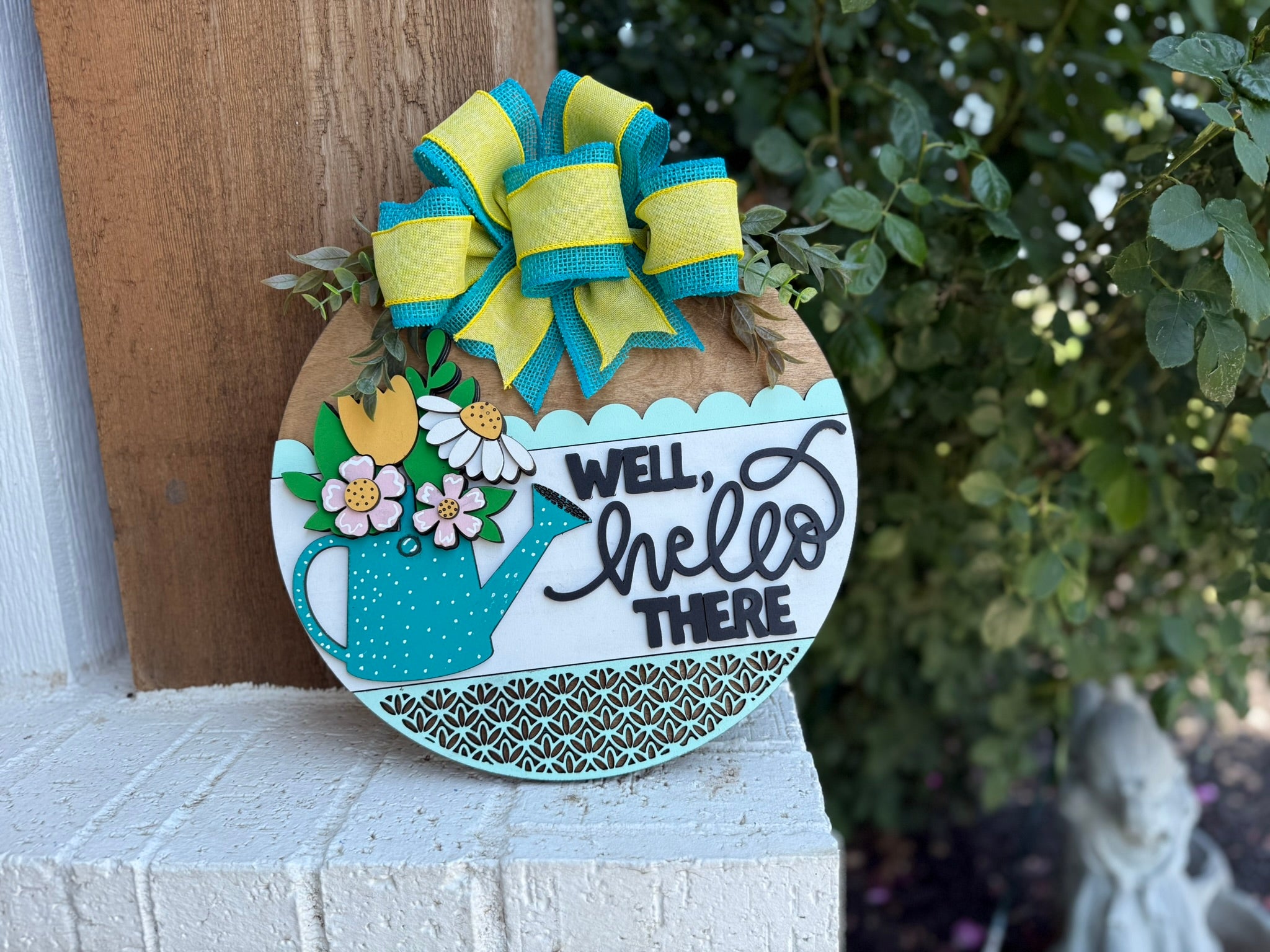 A round wooden sign decorated with a blue and yellow bow, a blue teapot with flowers, and the text “Well, hello there” hangs on a wall next to green bushes and a white brick surface.