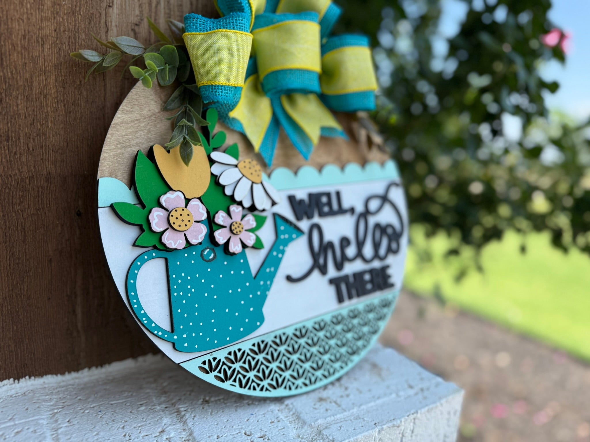 A round decorative wooden sign with a blue teapot, yellow and white flowers, and the words Well, hello there in script. It’s adorned with a green and yellow bow and greenery, hanging outdoors on a wooden surface.