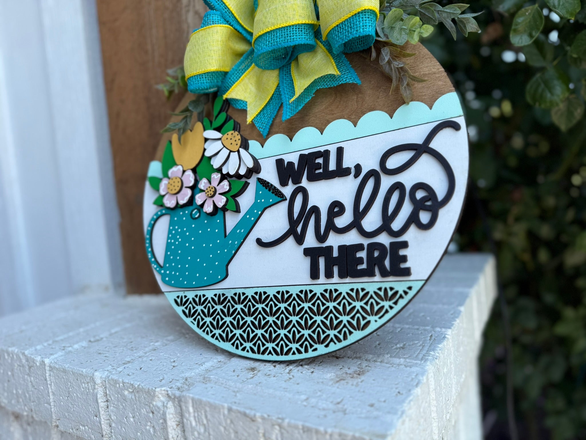 A decorative round sign with a teal watering can, flowers, and the text Well, hello there in black cursive letters, hanging on a brick surface with a yellow and teal bow above it.