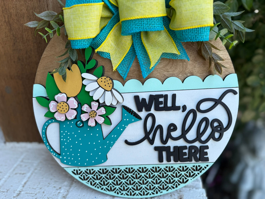 A decorative round wooden sign features a teal watering can with colorful flowers and leaves, a large yellow and teal bow, and the words Well, hello there in playful black script.