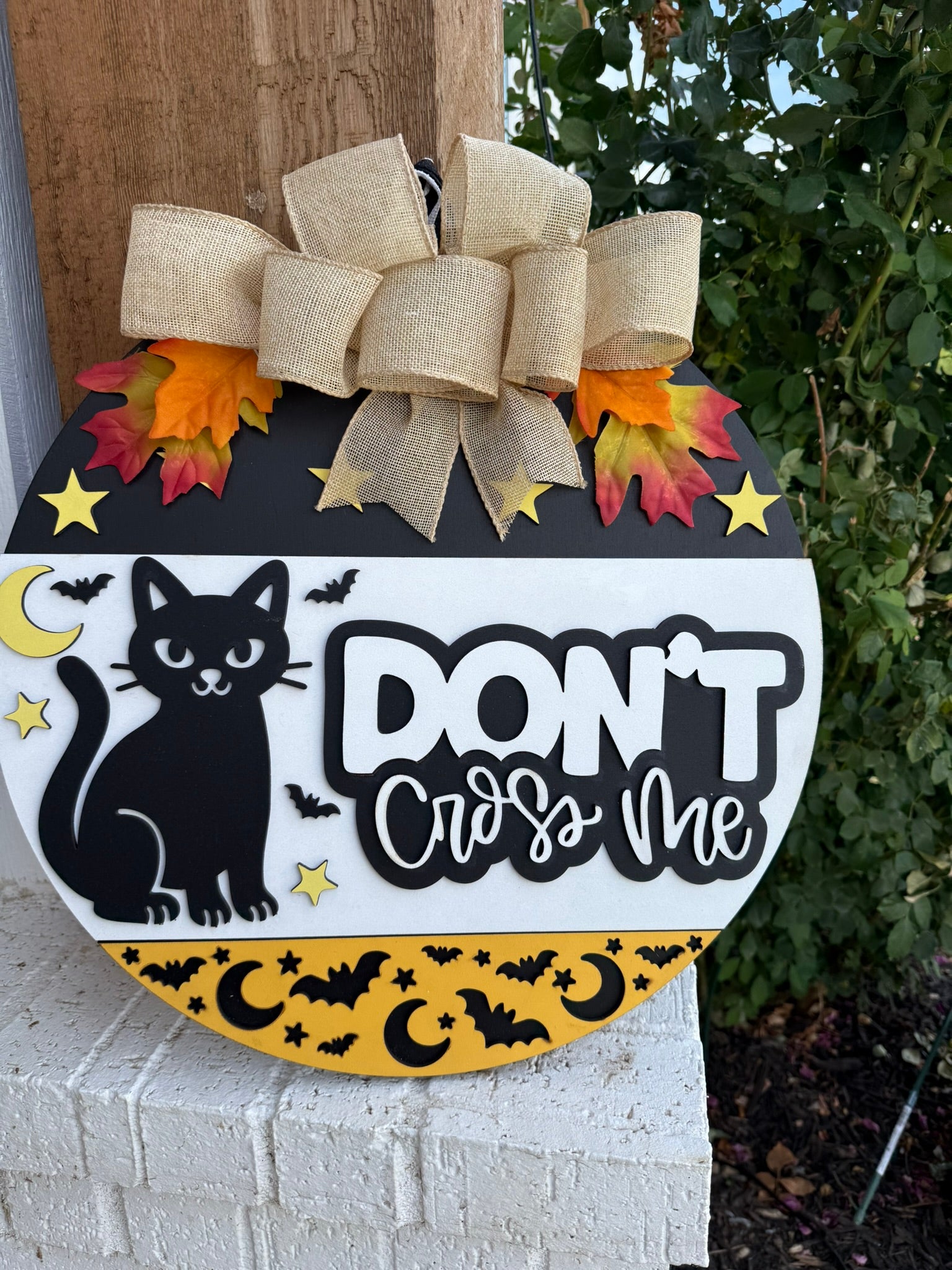 A round Halloween door sign features a black cat, yellow moons and bats, colorful leaves, a burlap bow, and the words Dont cross me in bold letters. The sign is propped against a brick wall near greenery.