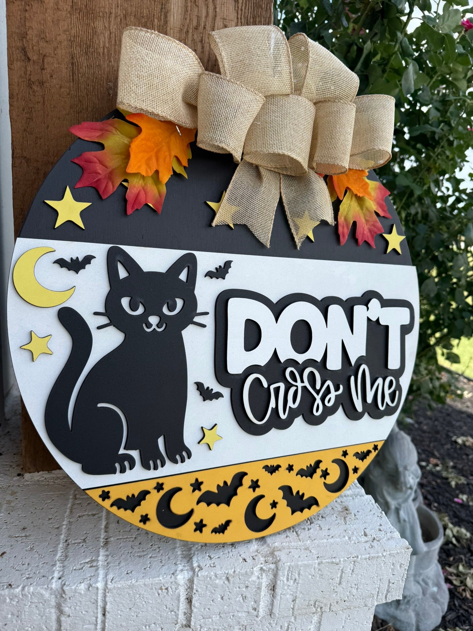 A round wooden sign decorated with a black cat, stars, crescent moons, bats, fall leaves, and a burlap bow. The sign reads “DONT Cross Me” in bold white and black letters.