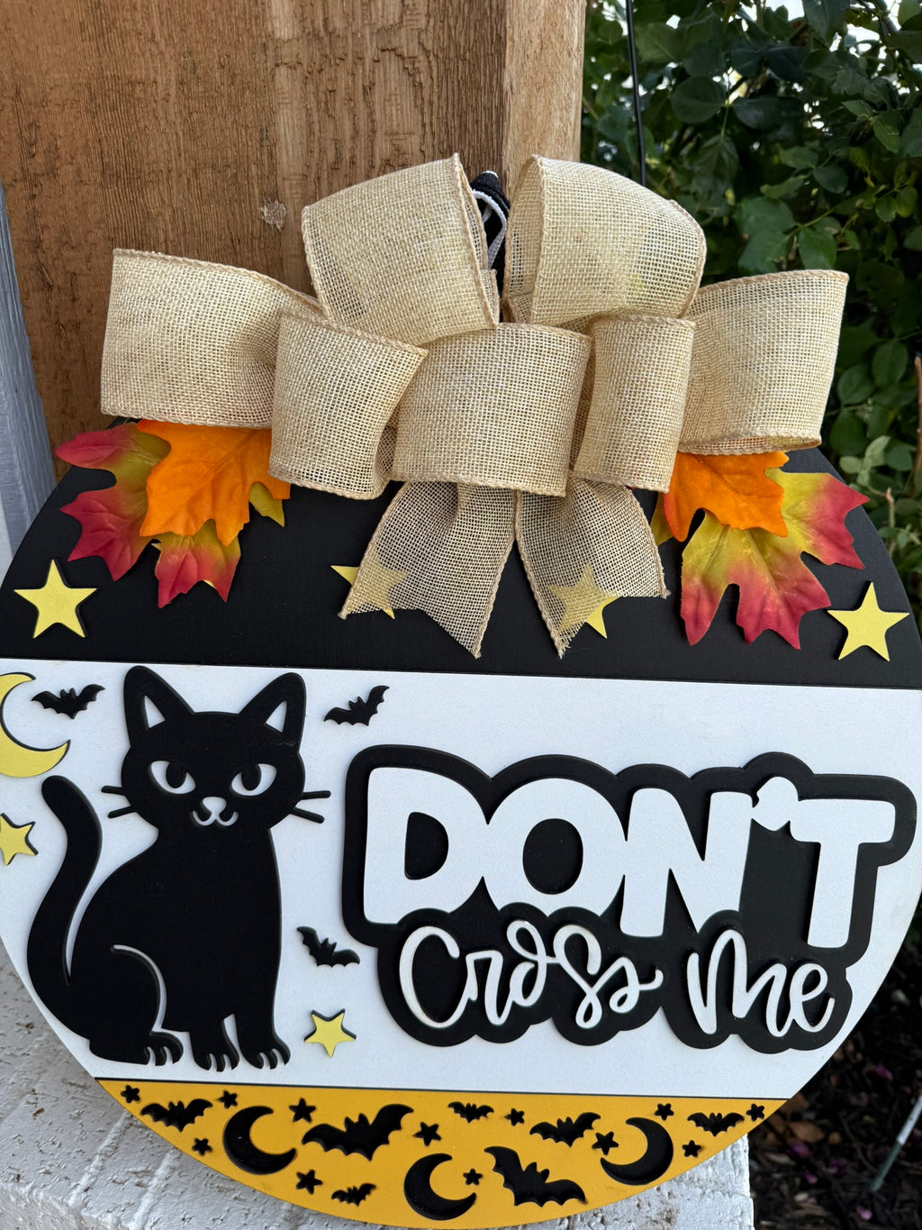 A round Halloween sign features a black cat, yellow stars, bats, and moons, with the words DONT cross me. The sign is decorated with a burlap bow and artificial autumn leaves at the top.