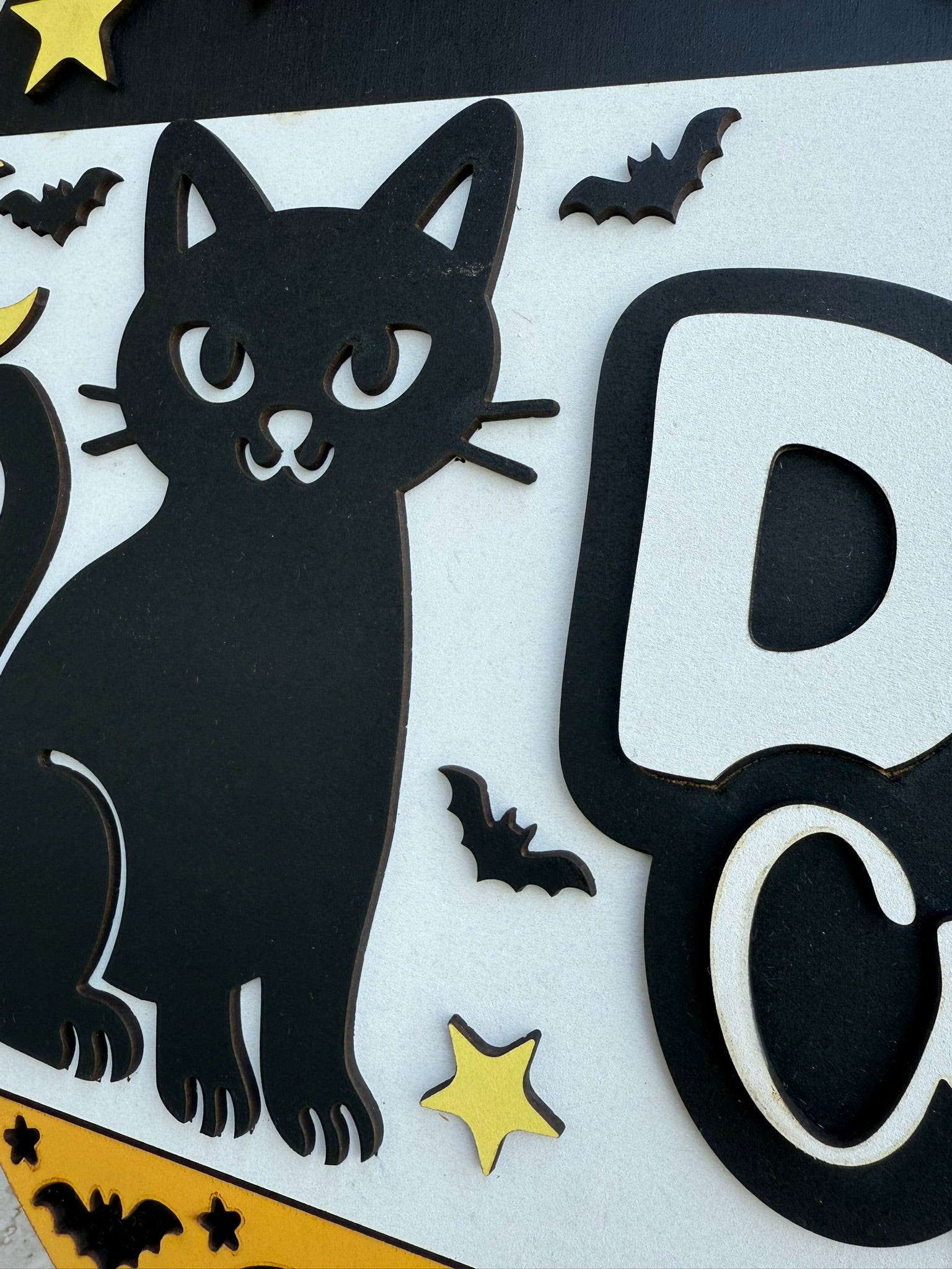 A decorative cutout of a black cat surrounded by small black bats and yellow stars on a white background. Part of a large letter D is visible on the right side of the image.