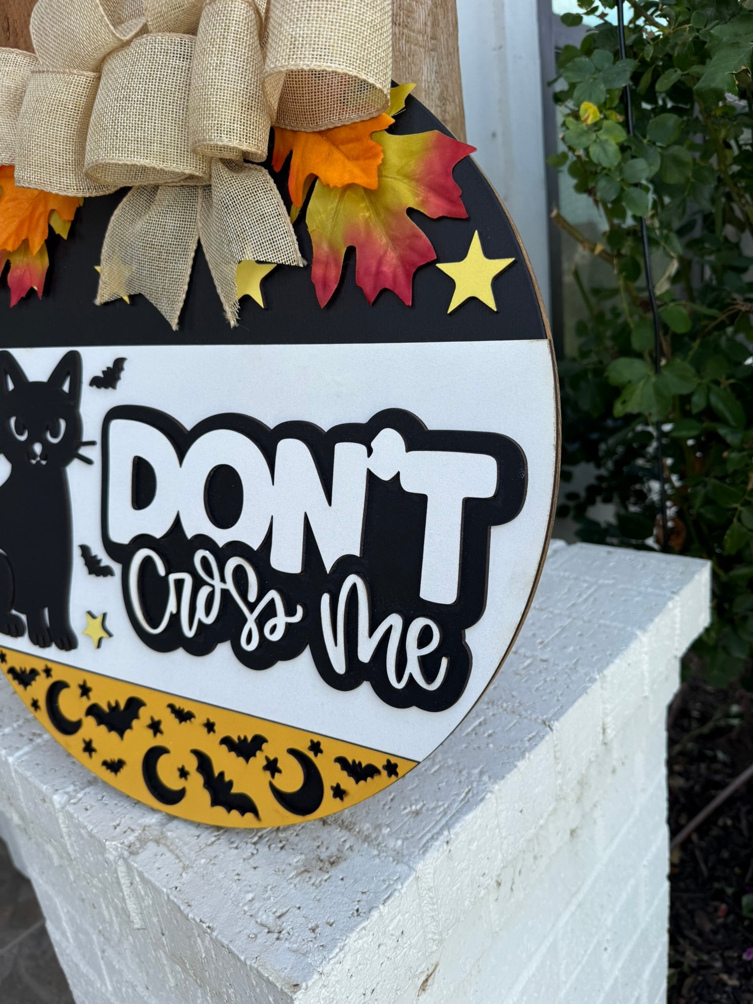 A round Halloween sign on a white brick surface reads DONT cross me with images of a black cat, bats, stars, and crescent moons, decorated with burlap ribbon and artificial autumn leaves.