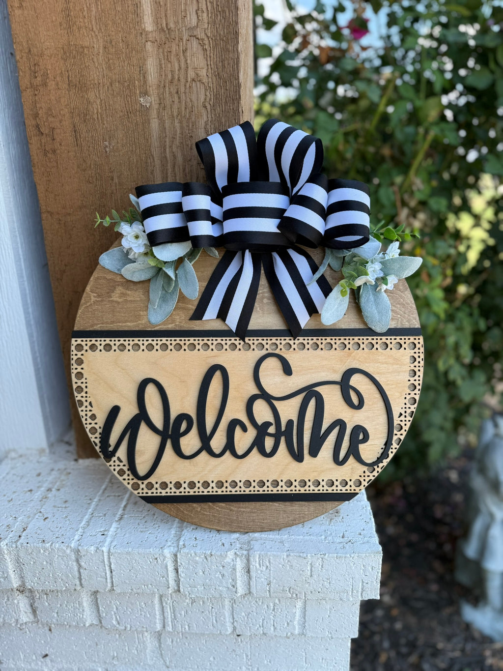 A round wooden sign with Welcome in script font, decorated with a black-and-white striped bow, faux greenery, and white flowers, hangs on a porch near a brick wall.