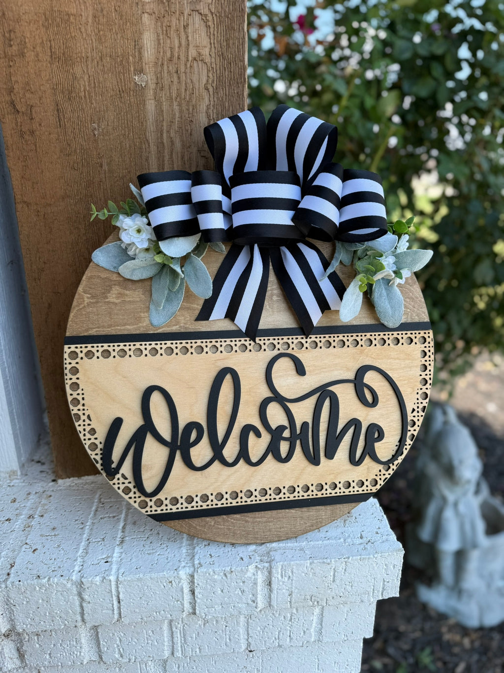A round wooden Welcome sign decorated with a black-and-white striped bow, white flowers, and greenery, hangs on a post next to a brick ledge, with greenery visible in the background.