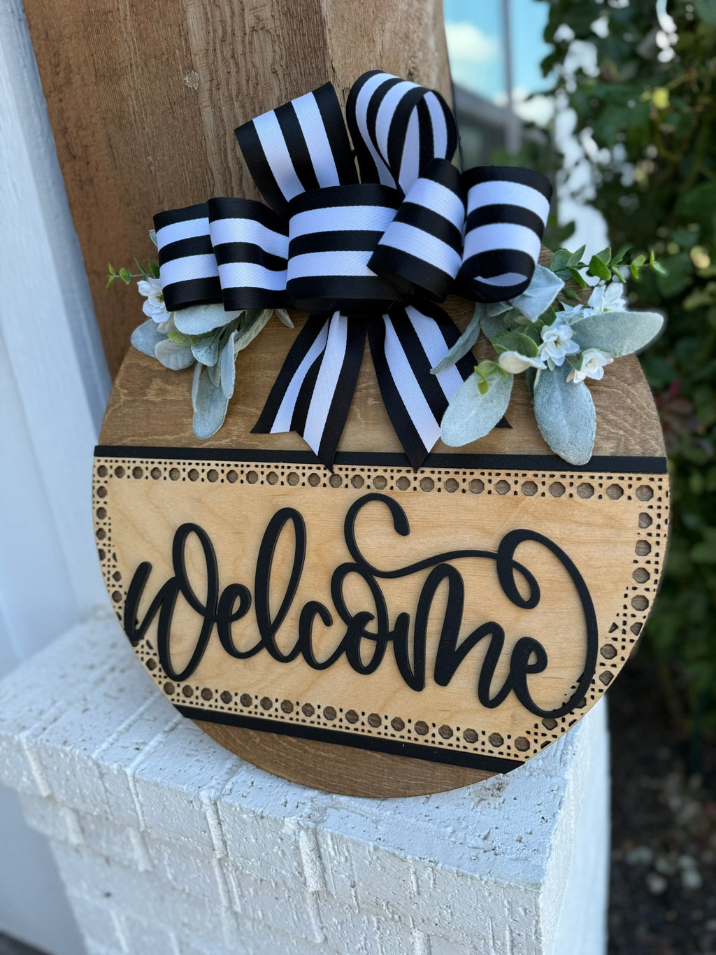 A round wooden sign with the word Welcome in black script, decorated with black-and-white striped ribbon and artificial greenery, is displayed on a white brick surface next to a wooden post.