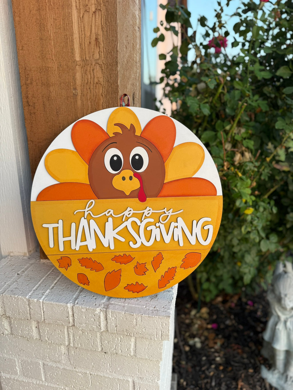 A round decorative sign with a cartoon turkey and colorful feathers reads Happy Thanksgiving in white and yellow letters, with autumn leaves at the bottom. It is hanging on a wall next to a garden.